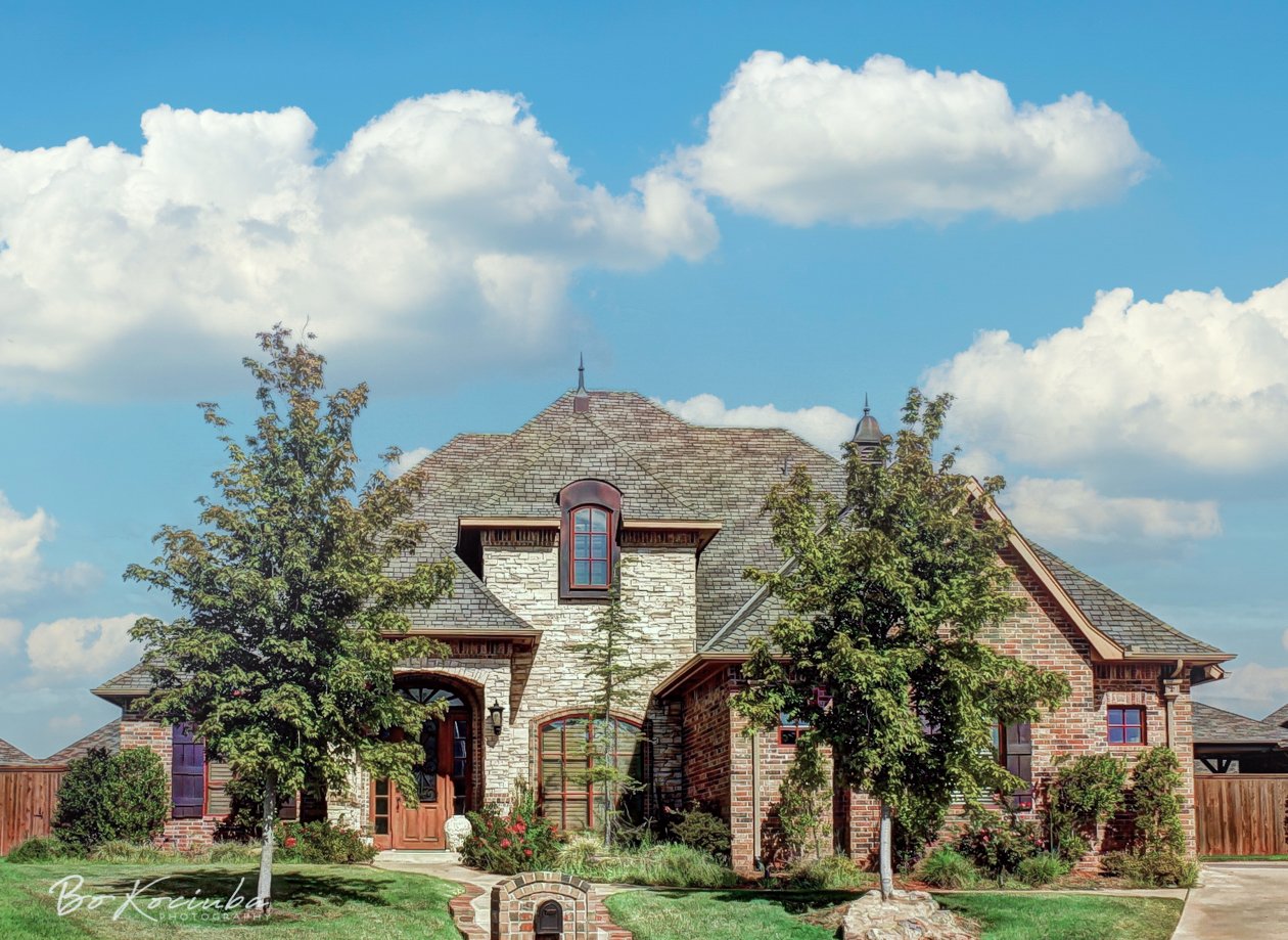 Brick home in Settlers Ridge, Yukon, Oklahoma, featuring a steeply pitched roof, mature trees, landscaped planting beds, and a well-kept front lawn.