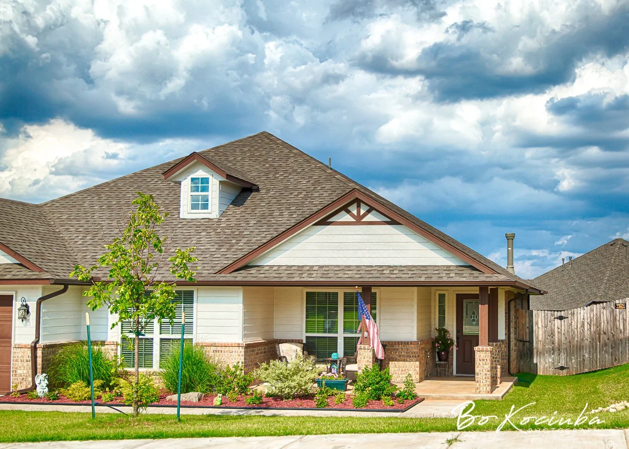 Brick and siding home in Chisholm Crossing, Yukon, Oklahoma, with a covered front porch, American flag, green lawn, and landscaped flower beds.