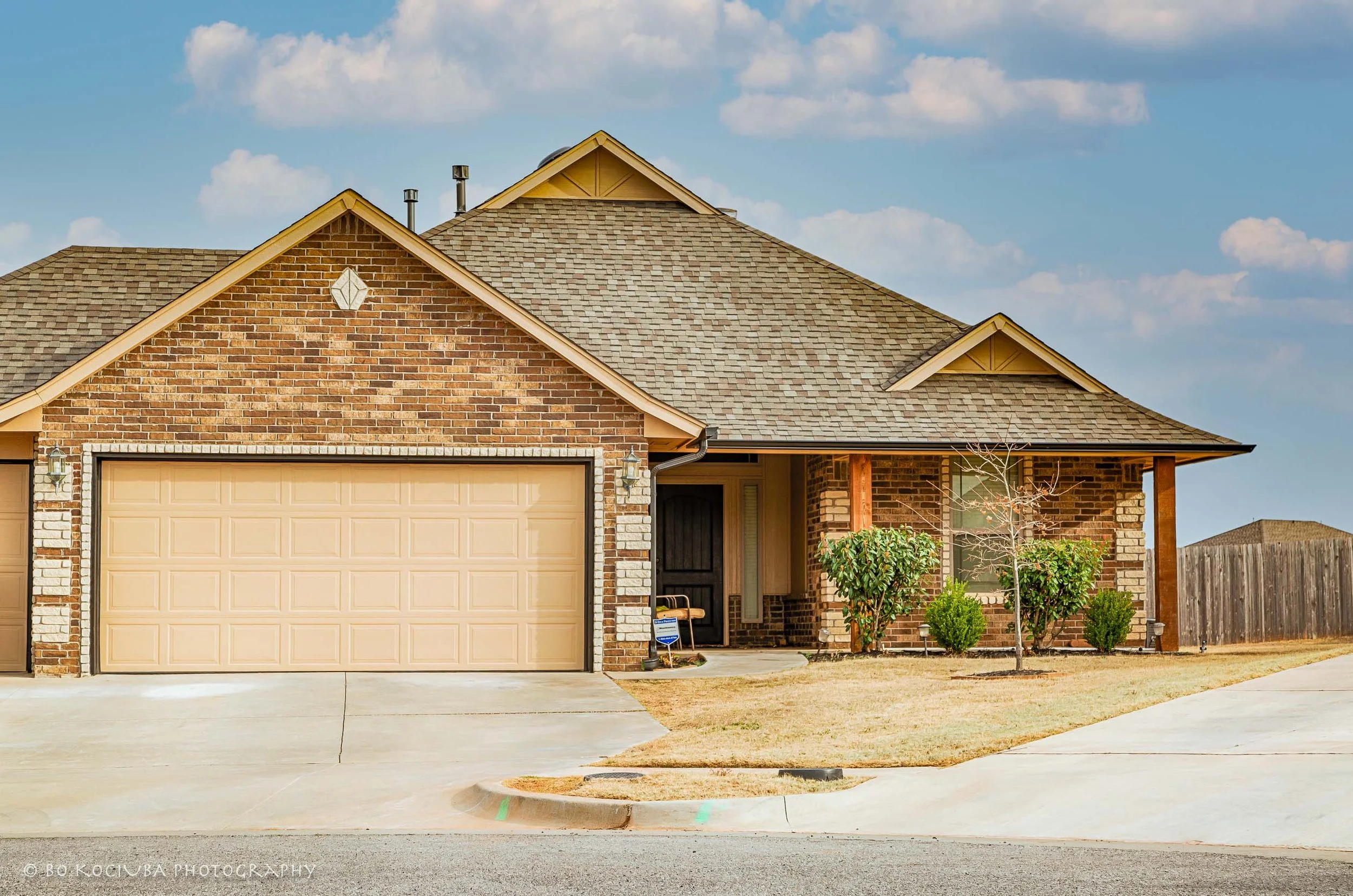 Brick home in Chisholm Crossing, Yukon, Oklahoma, with an attached two-car garage, covered front porch, concrete driveway, and young trees in the front yard.