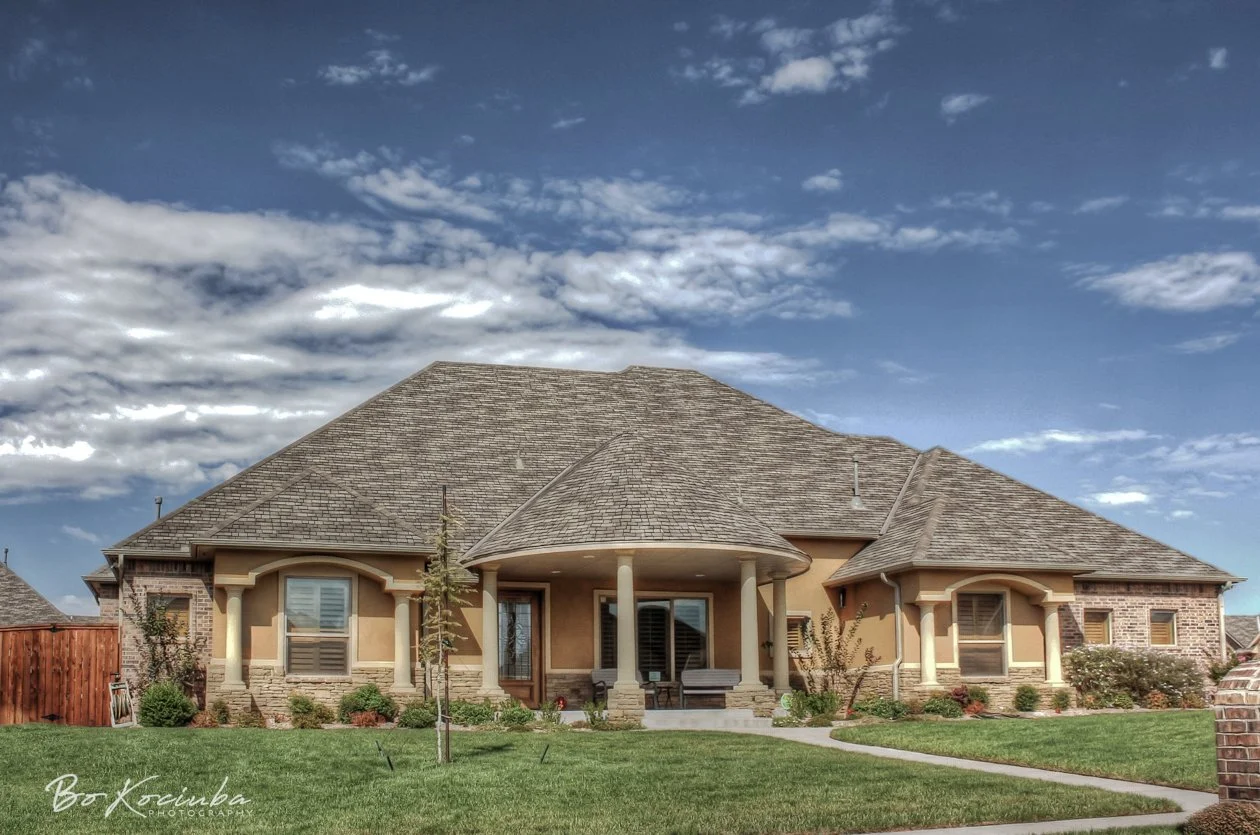 Spacious brick and stucco home in Settlers Ridge, Yukon, Oklahoma, featuring a curved covered porch, shuttered windows, and a landscaped front lawn.