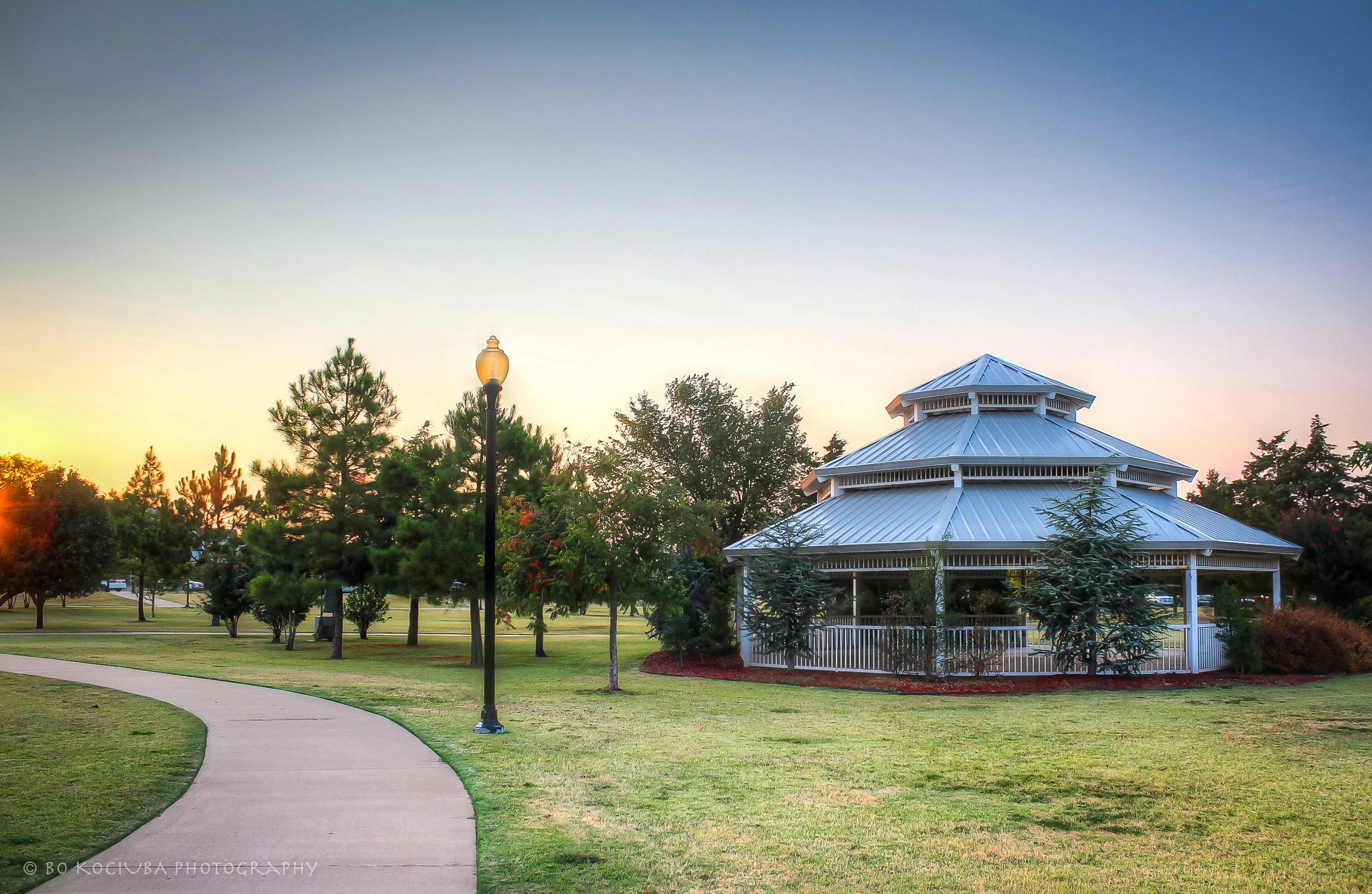 White two-tiered gazebo in a Mustang, Oklahoma, park at sunset, surrounded by trees, decorative streetlights, and a winding walking path.