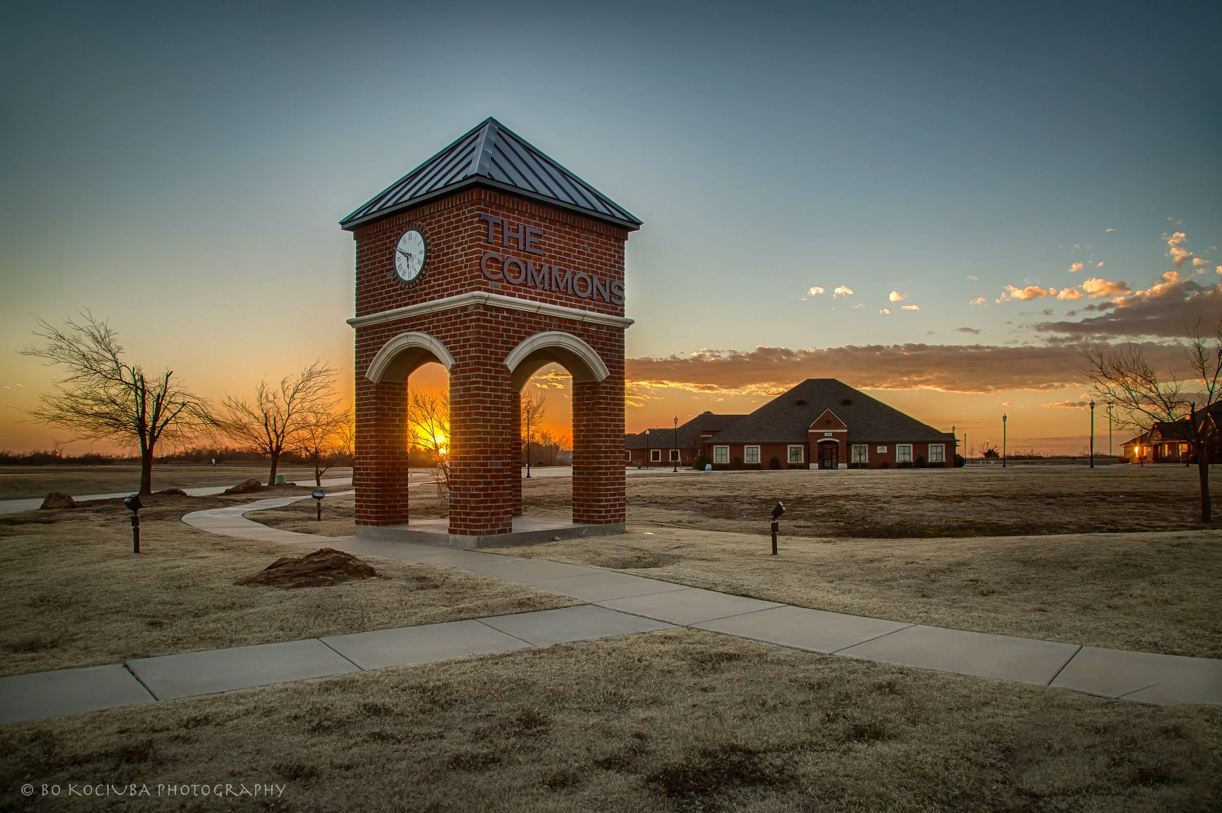 The Commons commercial area near Garth Brooks Boulevard in Yukon, Oklahoma, at sunset, showing brick homes and the intersection of The Commons Circle and Health Center Parkway.