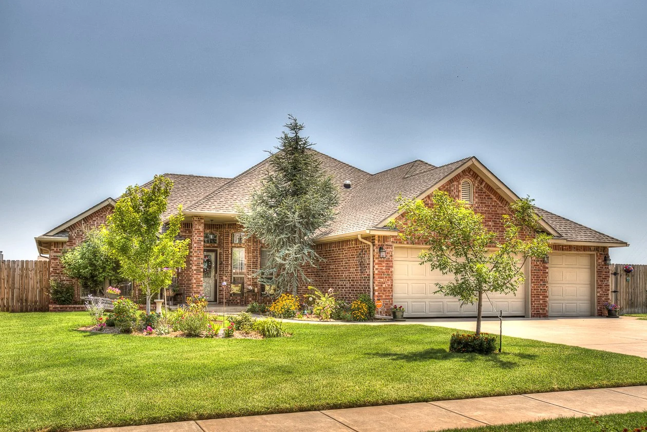 Single-story brick home in Hunters Hill, Mustang, Oklahoma, with a landscaped front yard, mature trees, flower beds, and an attached two-car garage.