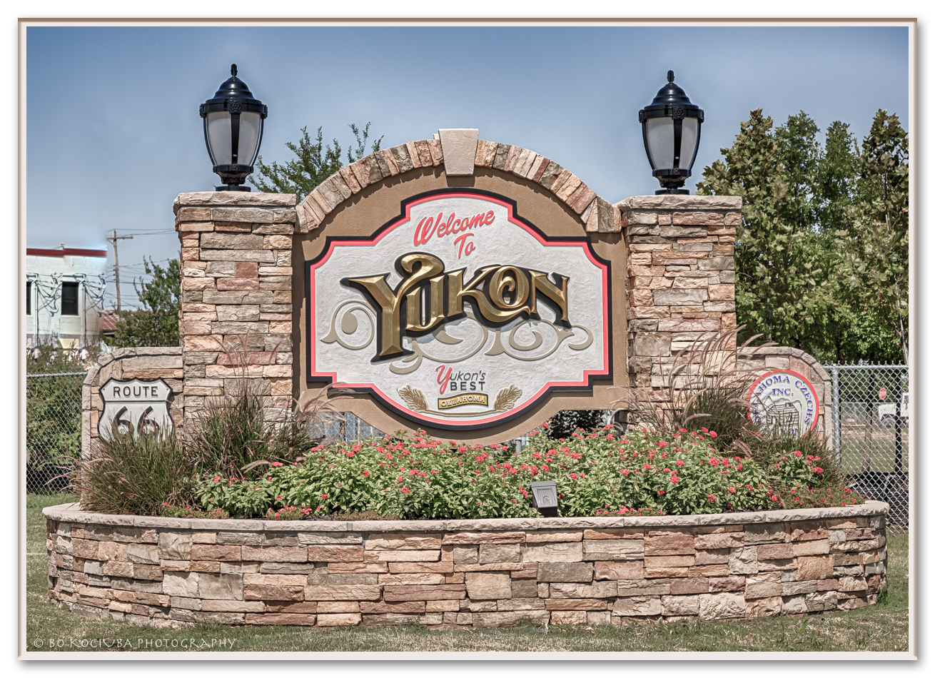 Welcome sign for Yukon, Oklahoma, made of stone with black lantern-style lights on top, surrounded by flowers and greenery.