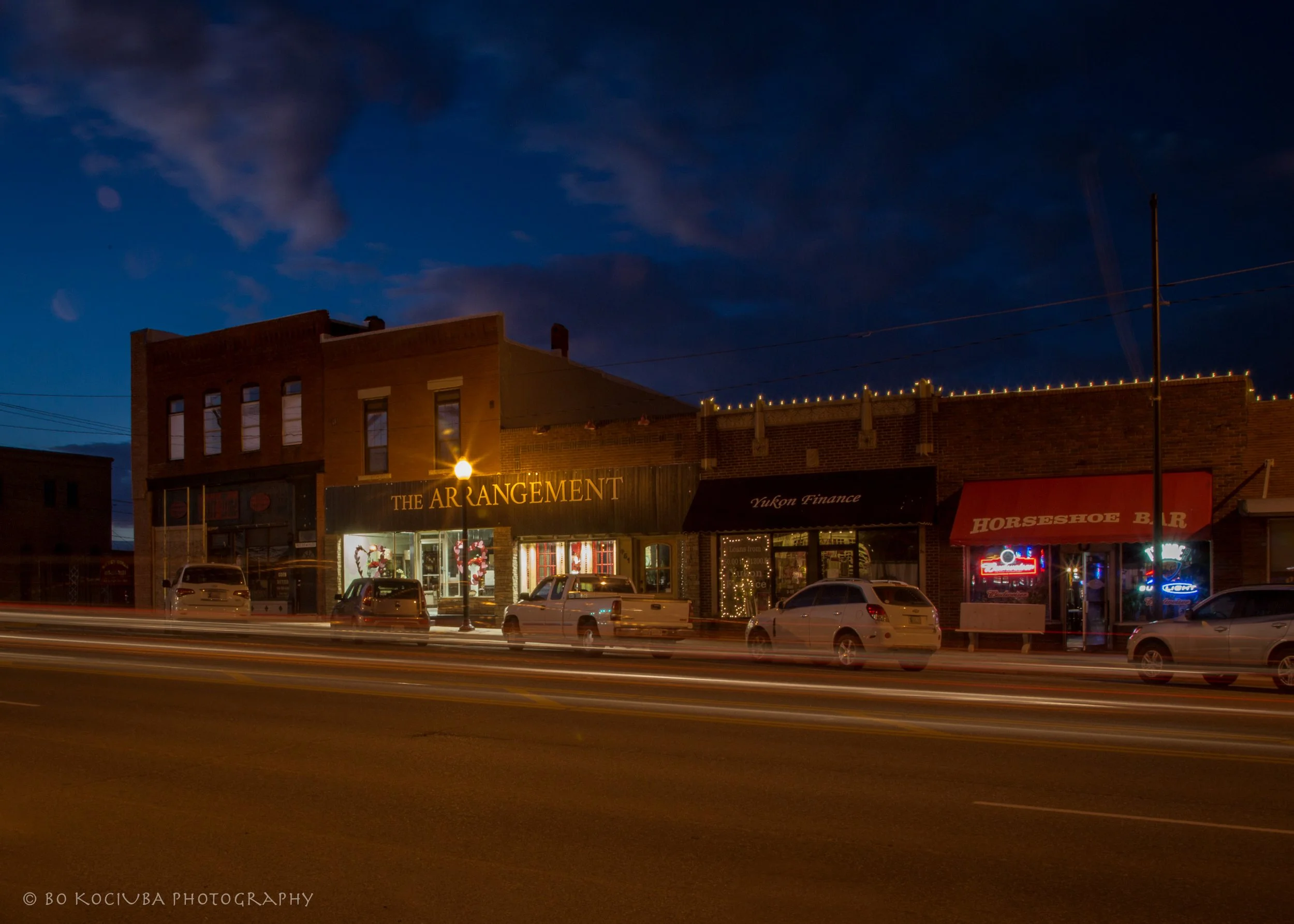 Brick storefronts along historic Main Street and Route 66 in downtown Yukon, Oklahoma, photographed at night with parked cars and passing traffic.