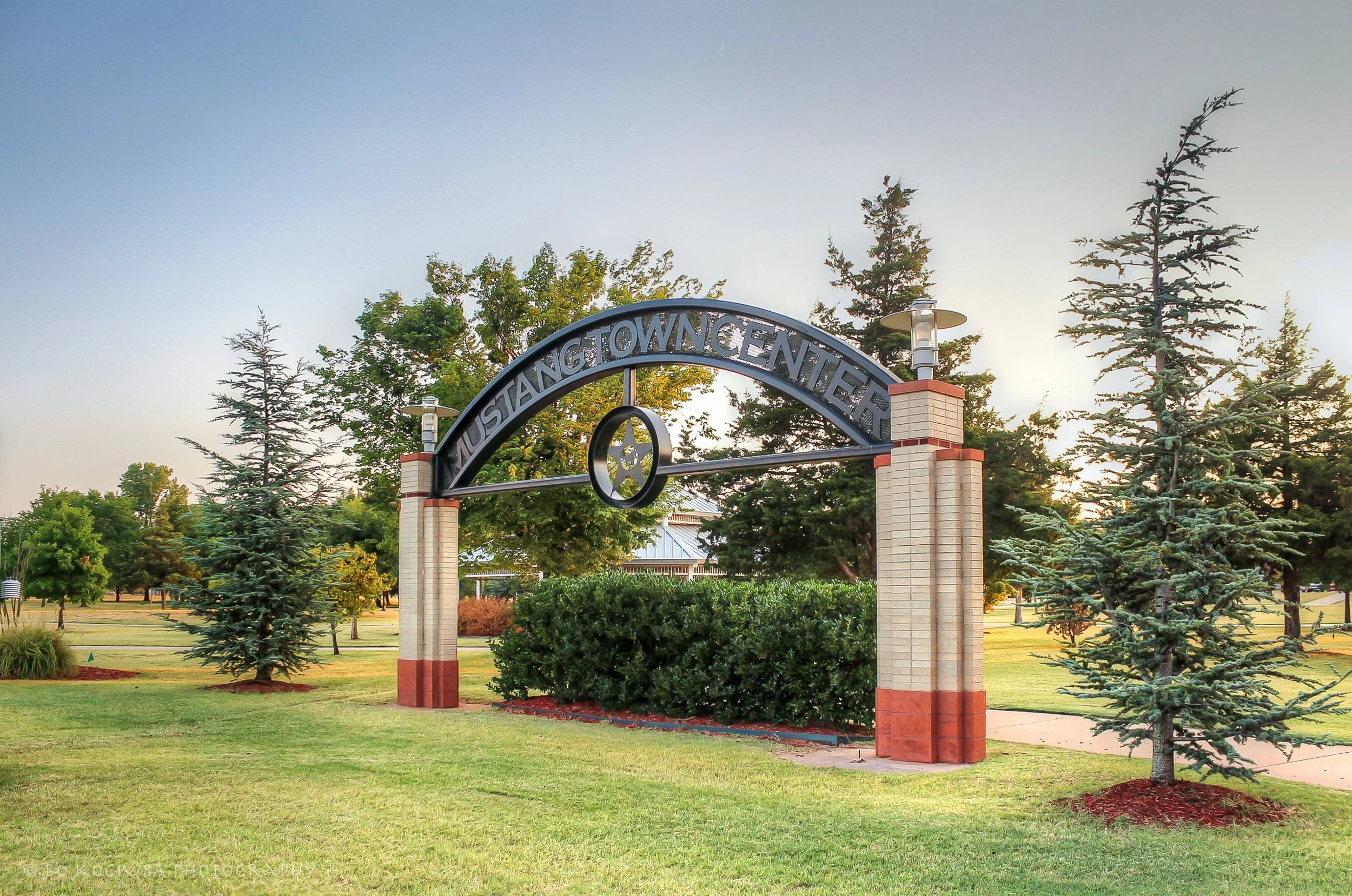 Mustang Town Center entrance arch in Mustang, Oklahoma, framed by brick columns, landscaped lawns, and mature trees.
