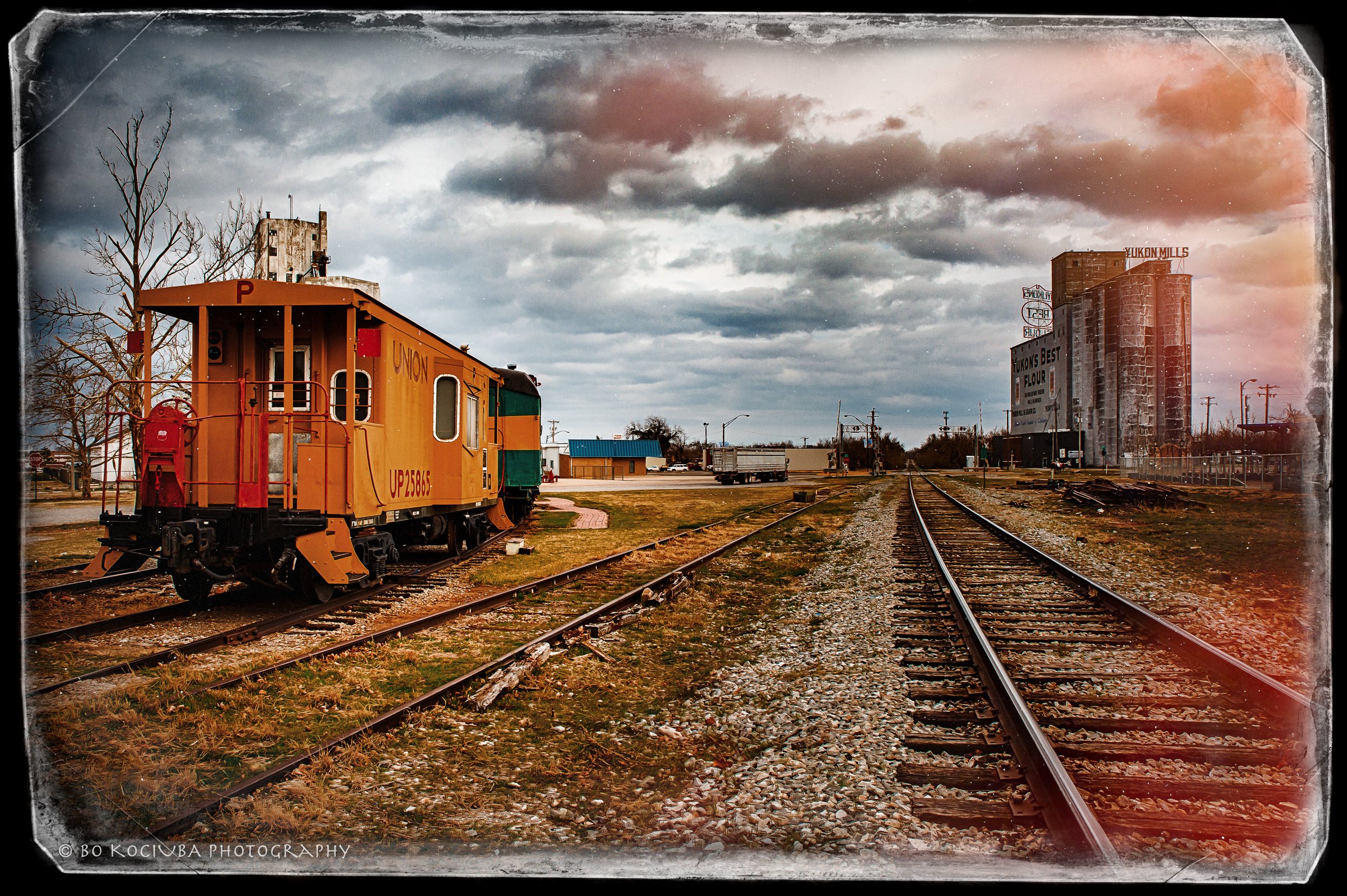 Historic orange Union Pacific locomotive on railroad tracks in Yukon, Oklahoma, with industrial buildings and grain elevators beneath a dramatic cloudy sky.