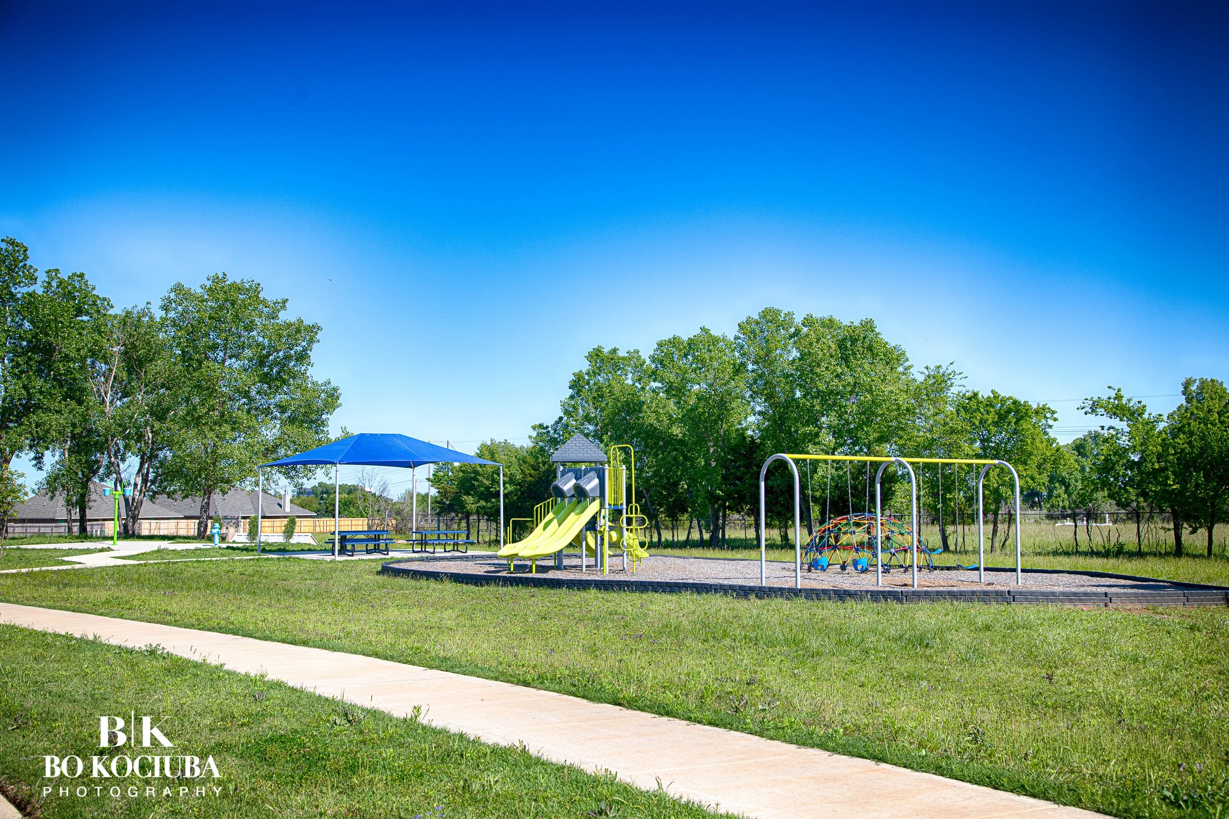 A playground with slides, swings, and a dome climbing structure surrounded by green trees and grass under a clear blue sky.
