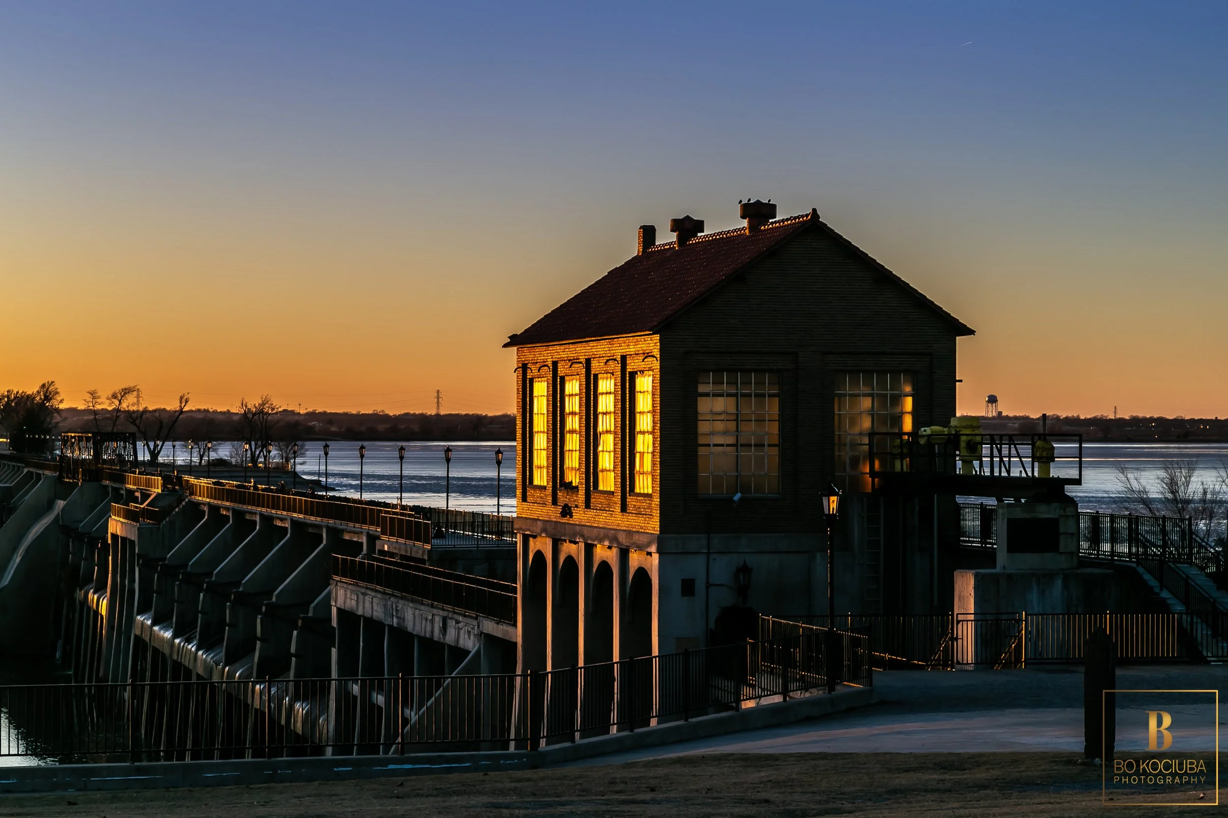 Historic brick building at Lake Overholser Dam near Yukon, Oklahoma, illuminated by sunset light, with the dam structure and lake visible beside it.