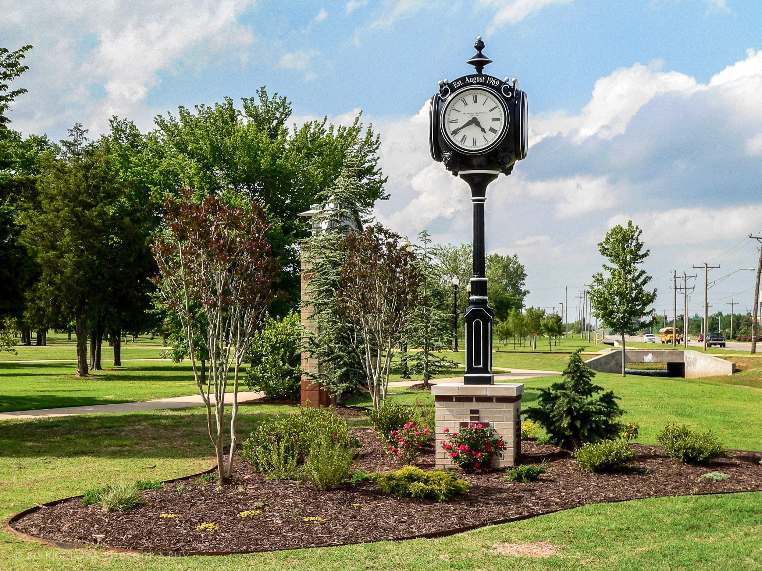 Decorative black street clock with Roman numerals in a landscaped public park in Mustang, Oklahoma, surrounded by trees, flowering plants, and a walking path.