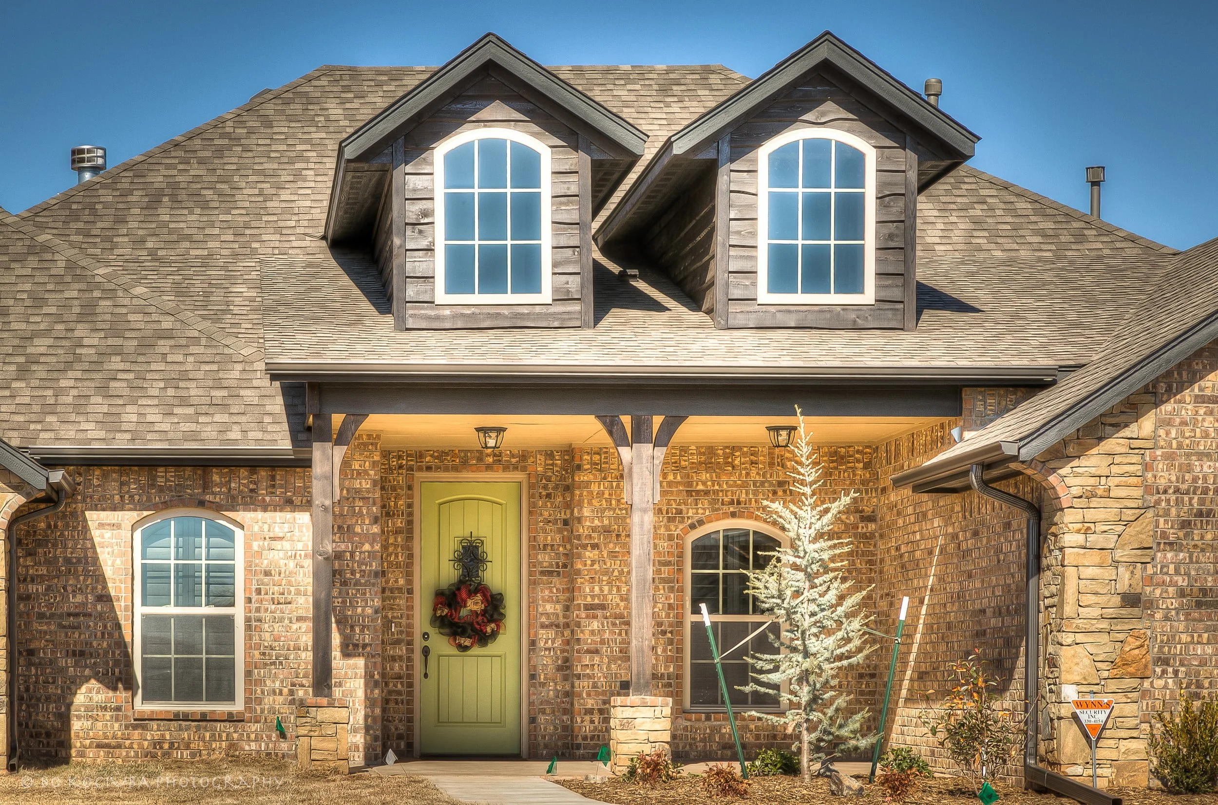 Brick home in Chisholm Run, Yukon, Oklahoma, with two dormer windows, a green front door decorated with a wreath, and a covered entry with stone columns.