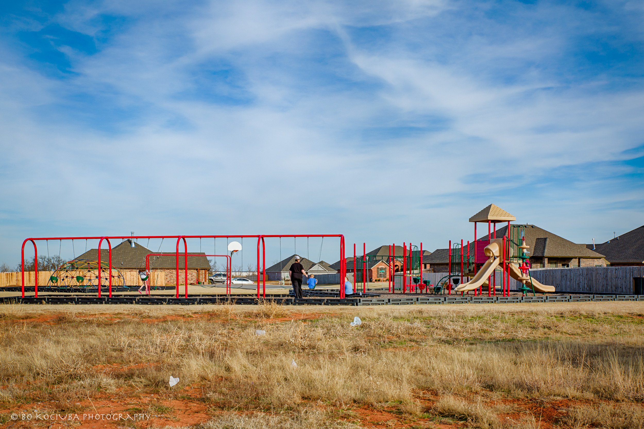 A playground with red swings, a climbing dome, a slide, and a small tower, surrounded by a residential neighborhood with houses and a wooden fence, under a partly cloudy blue sky.