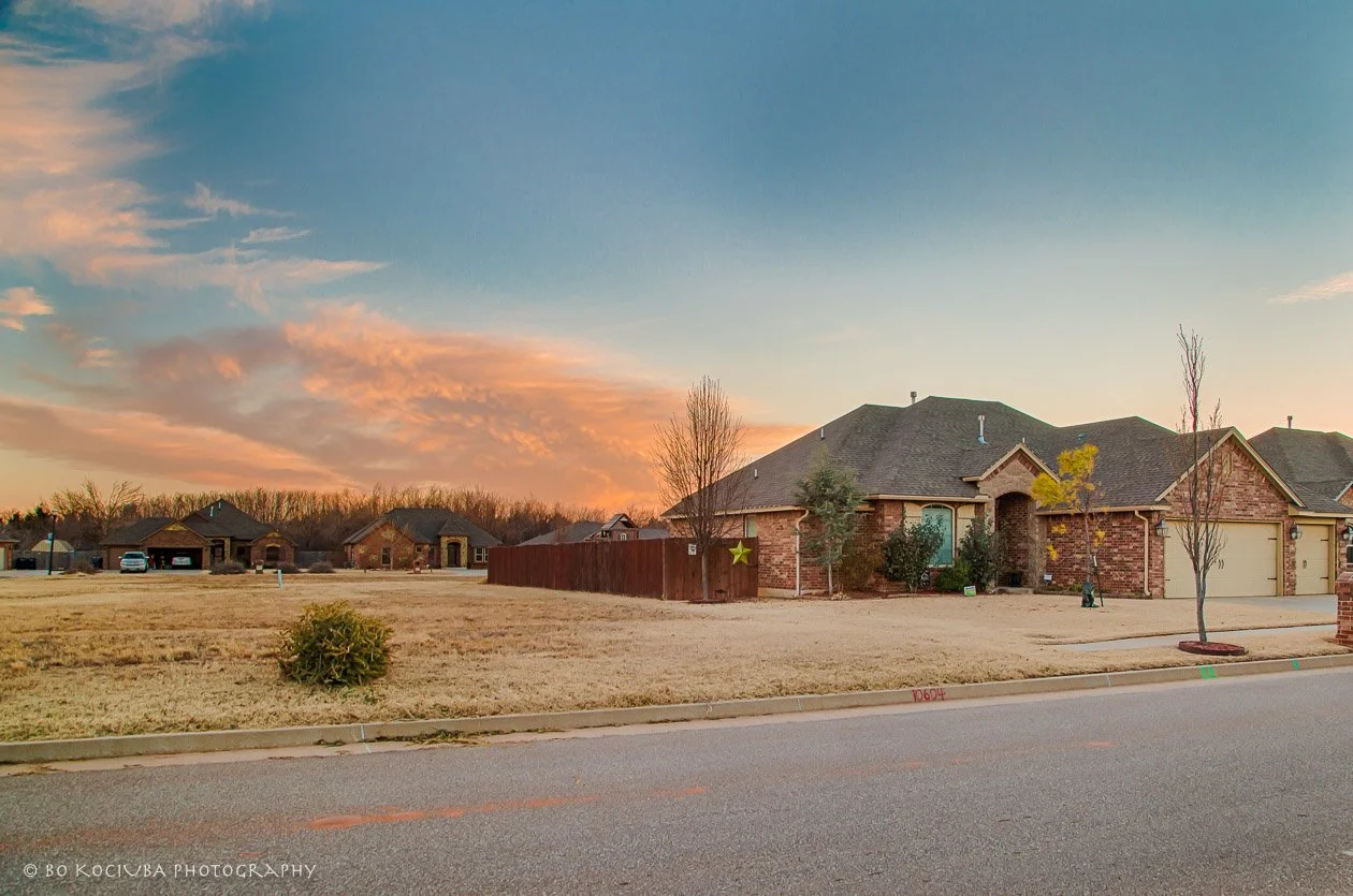 Residential street in Sabram Estates, Yukon, Oklahoma, lined with brick homes, landscaped yards, and trees beneath a colorful sunset sky.