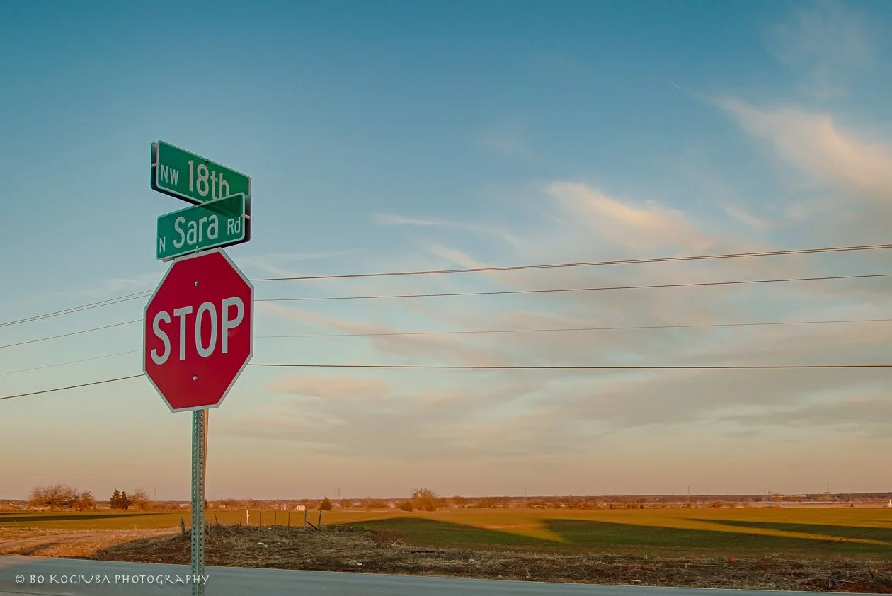 Intersection of NW 18th Street and Sara Road near Sabram Estates in Yukon, Oklahoma, with street signs, a stop sign, and open fields at sunset.