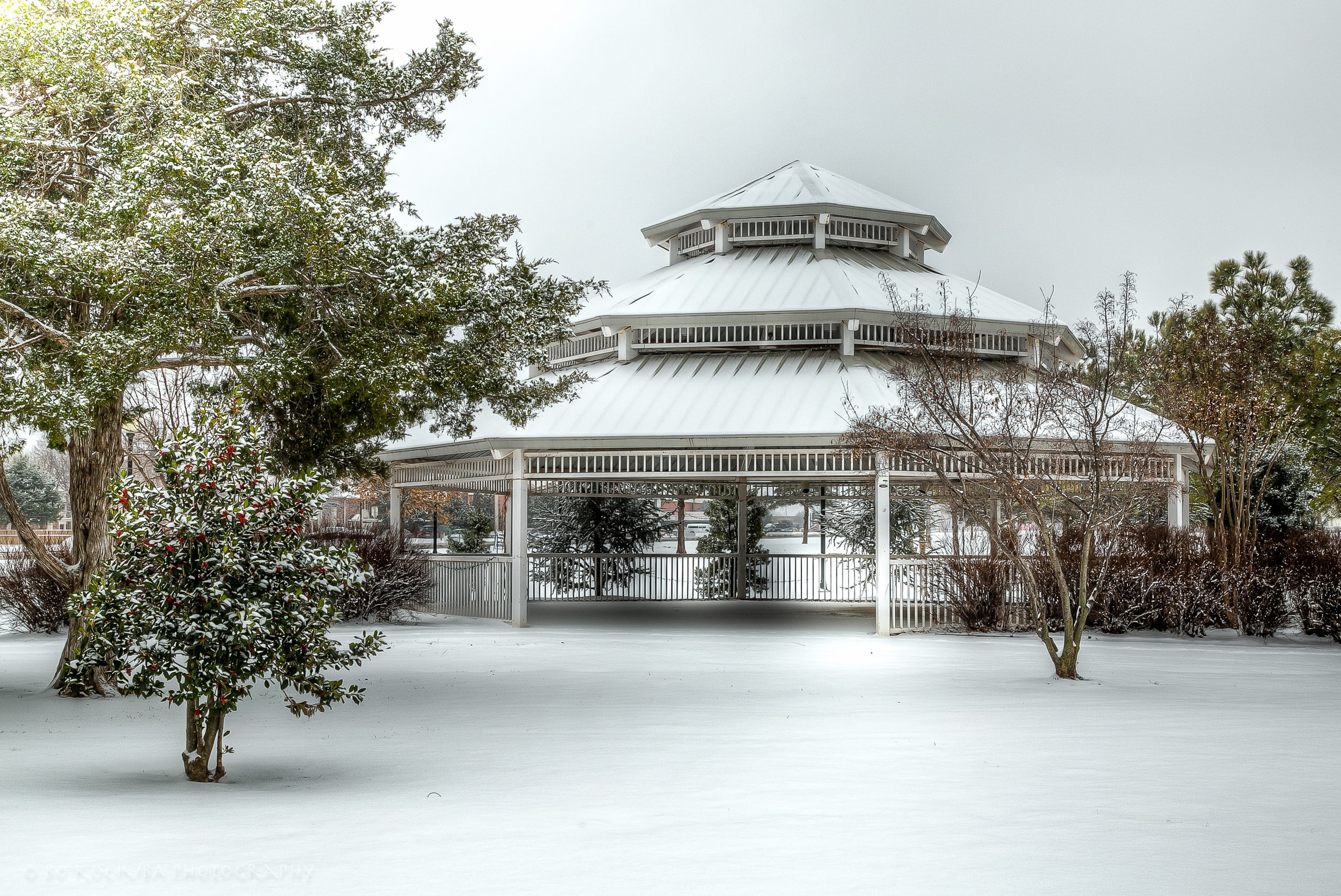 Two-tiered white park gazebo surrounded by snow-covered trees and grounds during winter in Mustang, Oklahoma.