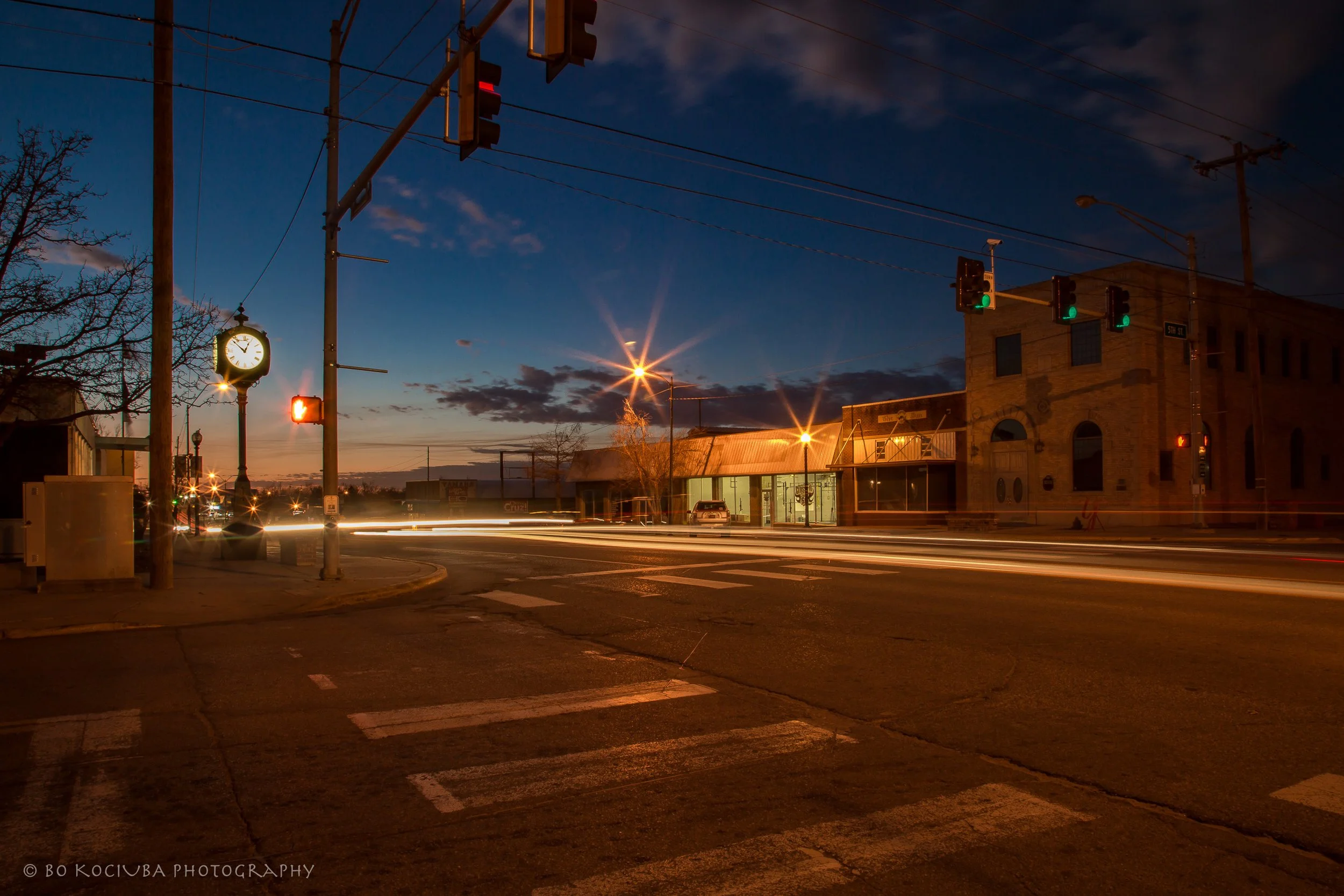 Historic Main Street along Route 66 in Yukon, Oklahoma, at dusk, with a decorative street clock, traffic signals, and illuminated downtown buildings.