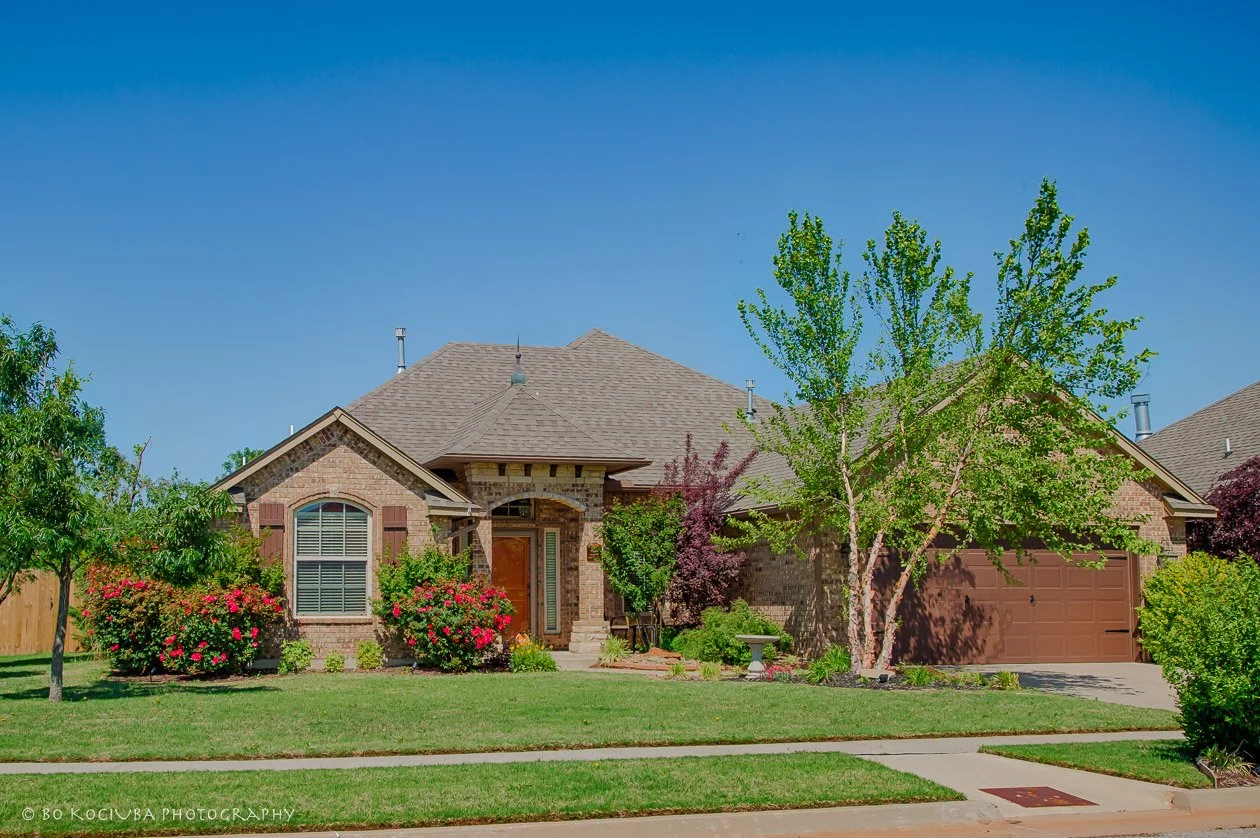 Single-story brick home in Sabram Estates, Yukon, Oklahoma, featuring a landscaped front lawn, flowering shrubs, mature trees, and an attached garage.