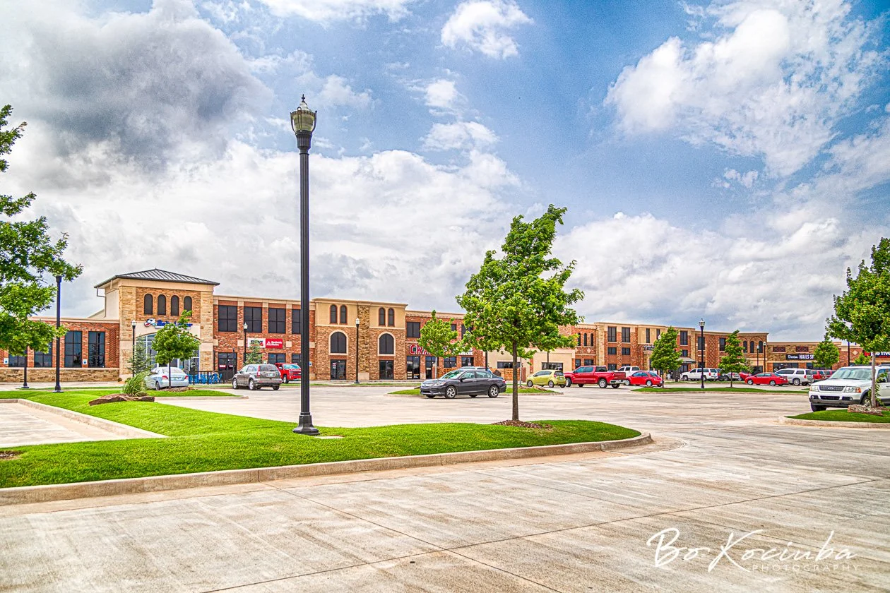 Shopping Plaze in Yukon, Oklahoma, with landscaped grounds, trees, parking areas, and business buildings beneath a partly cloudy sky.
