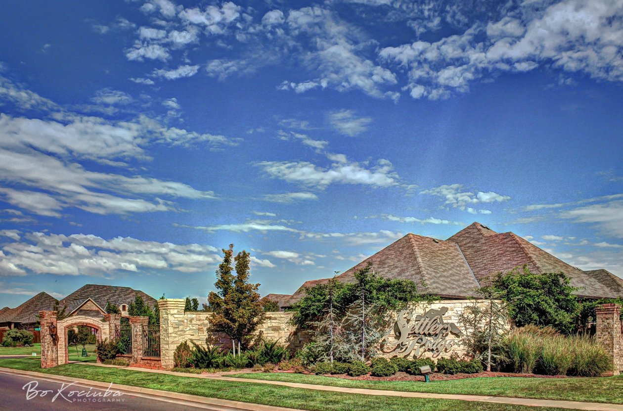 Landscaped gated entrance to Settlers Ridge in Yukon, Oklahoma, featuring the neighborhood entrance monument, mature trees, and brick homes beyond the gate.