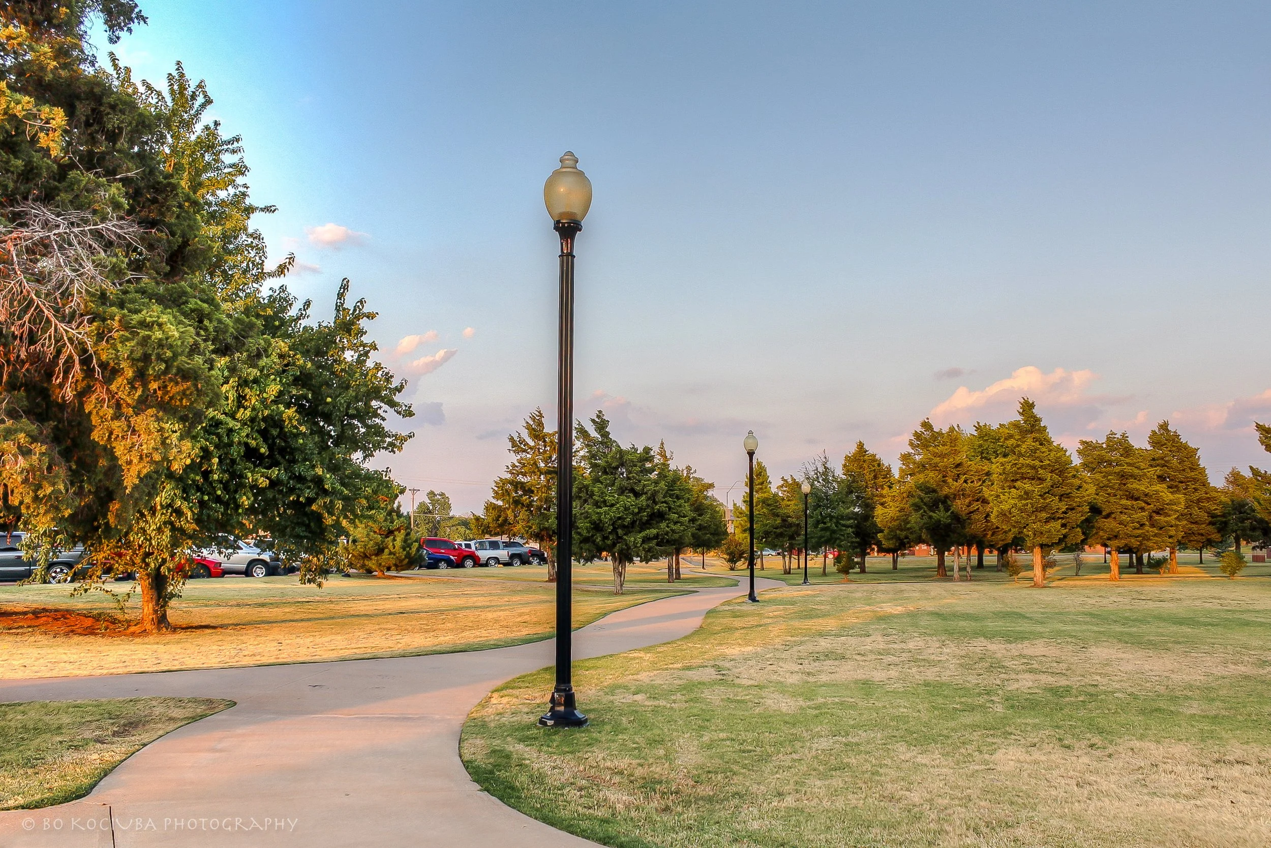 Paved walking trail through a park in Mustang, Oklahoma, bordered by trees, grassy areas, and decorative streetlights in the late afternoon.