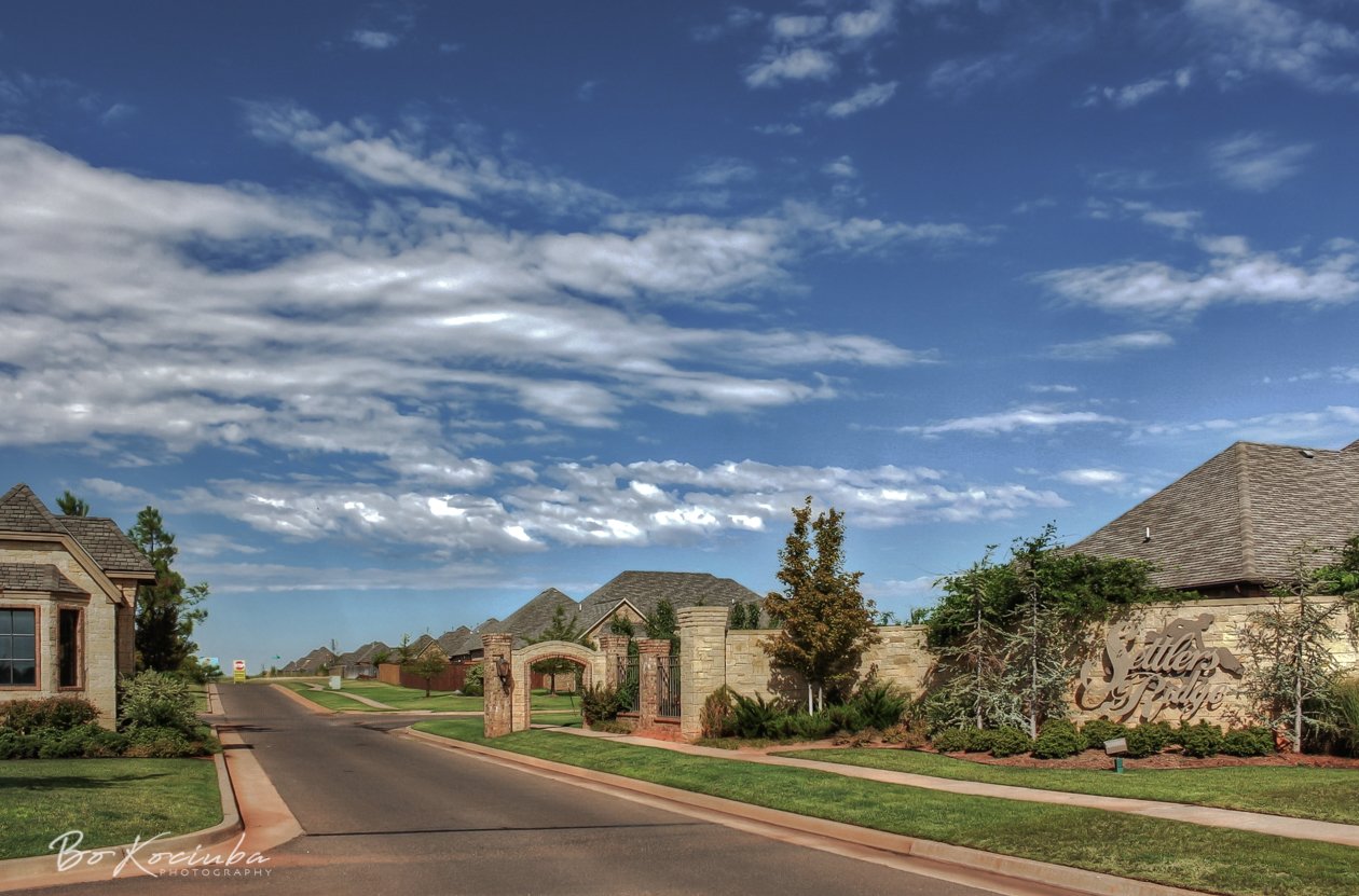 Landscaped entrance to the gated Settlers Ridge neighborhood in Yukon, Oklahoma, featuring brick and stone entrance monuments, a residential street, and homes beyond the entrance.