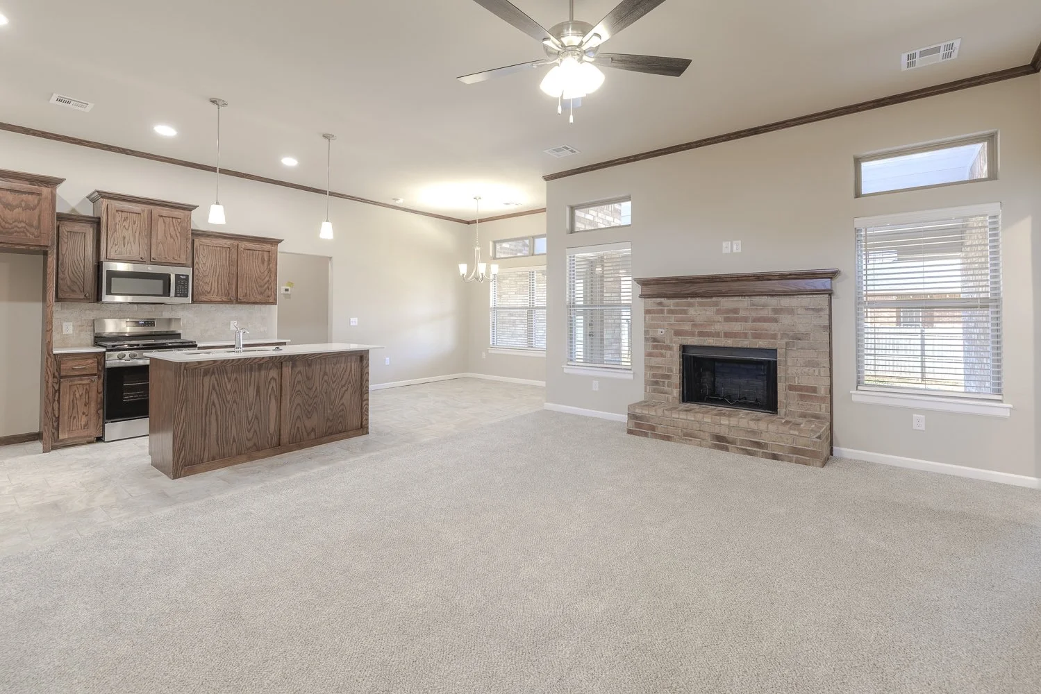 Empty living room with beige carpet, brick fireplace, large windows with white blinds, open kitchen with wooden cabinets, island, and stainless steel appliances, ceiling fan, and natural light.