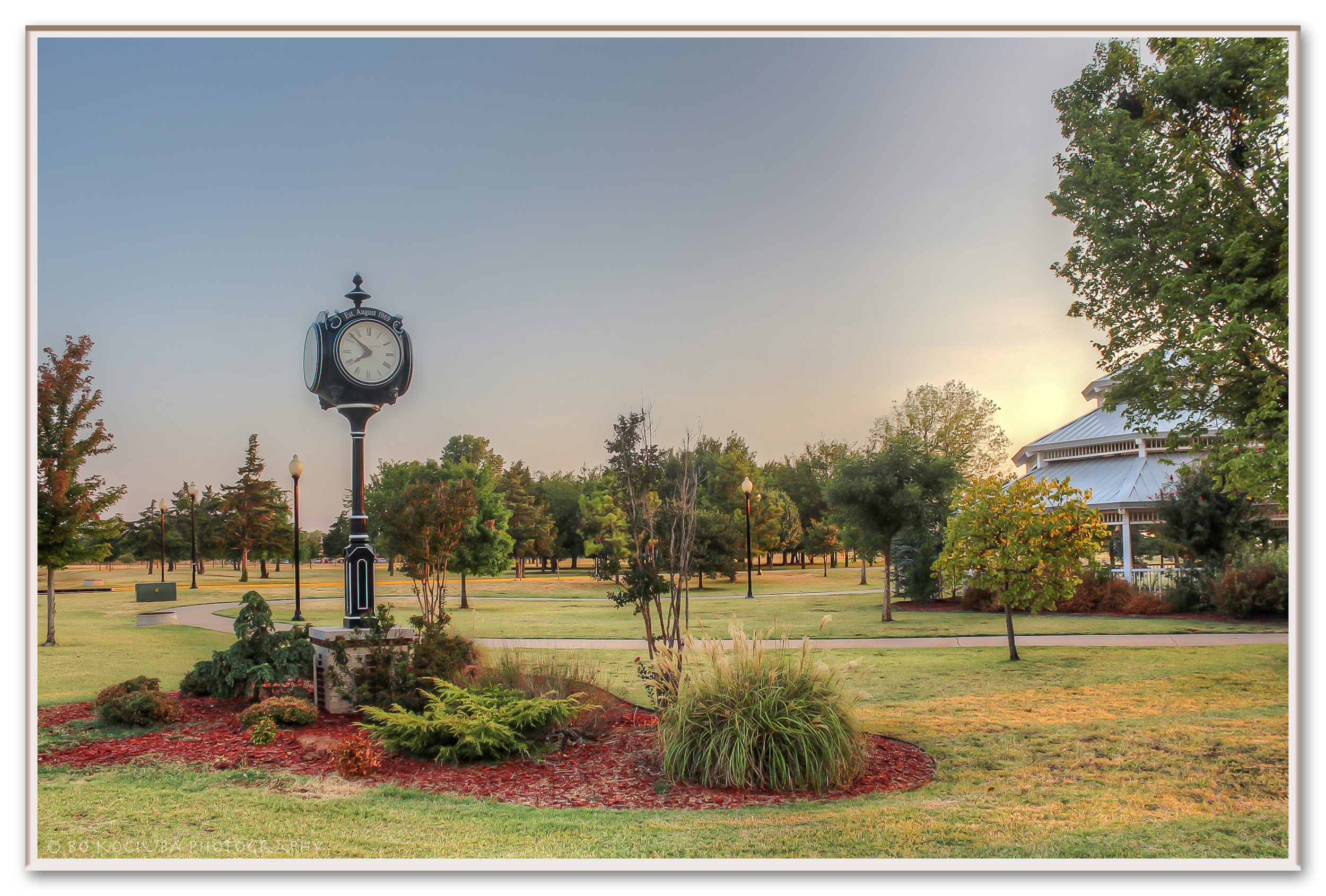 A park with a vintage clock on a black pole, landscaped with bushes and red mulch, green grass, tall trees, a gazebo, and lampposts, during sunset.
