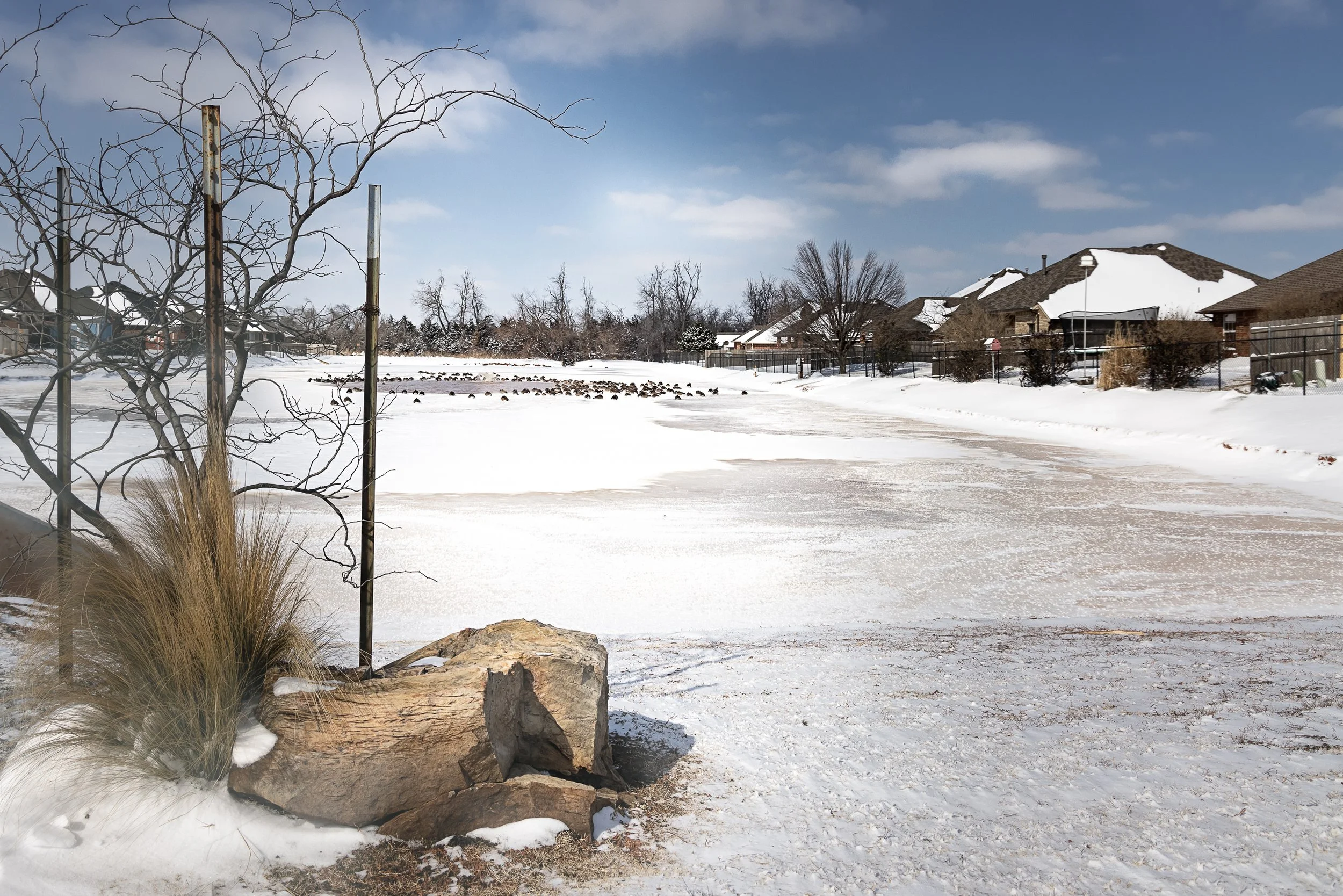 Snow-covered shoreline of the Hunters Hill neighborhood pond in Mustang, Oklahoma, with winter trees, ornamental rocks, and homes across the water.