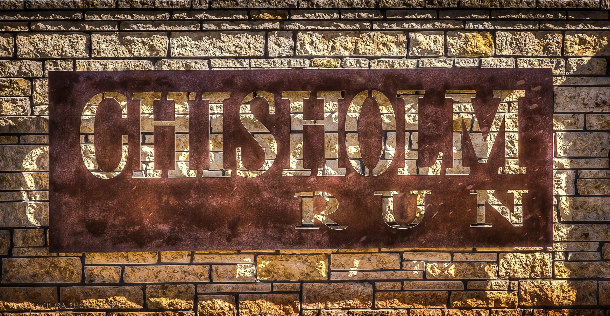 Chisholm Run neighborhood entrance sign mounted on a brick wall in the Yukon, Oklahoma, area, featuring rustic metal lettering and decorative stonework.
