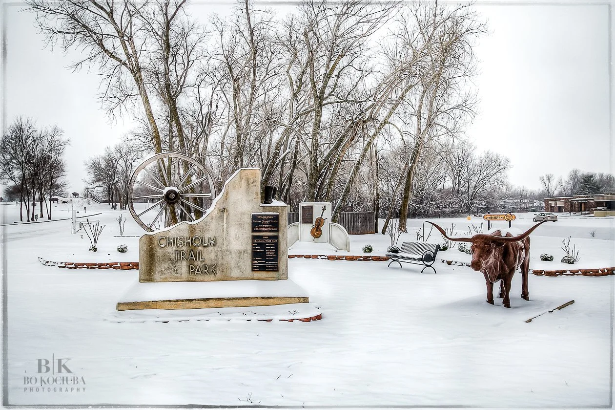 Chisholm Trail Park in Yukon, Oklahoma, during winter, with snow covering the park entrance, historic wagon wheel display, and outdoor sculptures.