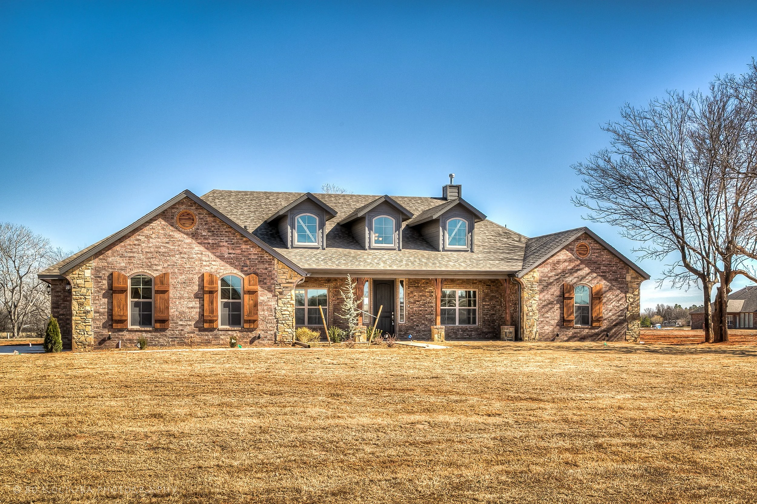 Brick home in Chisholm Run, Yukon, Oklahoma, featuring three dormer windows, a wide front lawn, and mature trees beneath a clear blue sky.