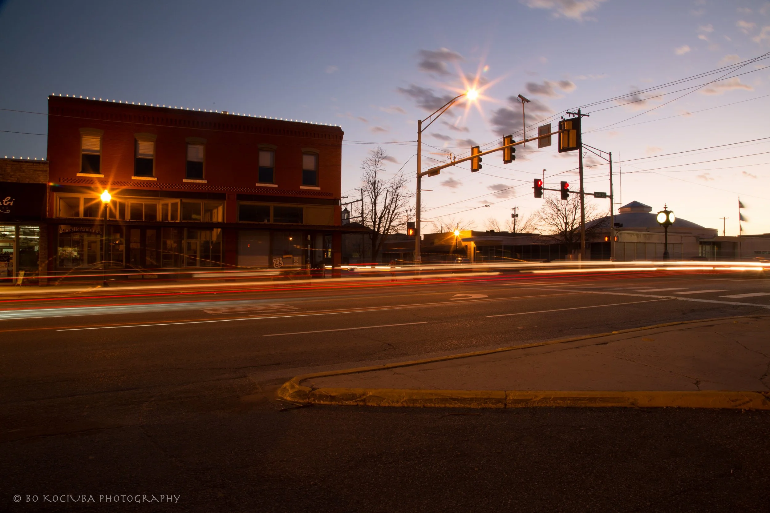 Downtown Yukon, Oklahoma, along historic Route 66 at dusk, showing Main Street buildings, a traffic intersection, and light trails from passing cars.