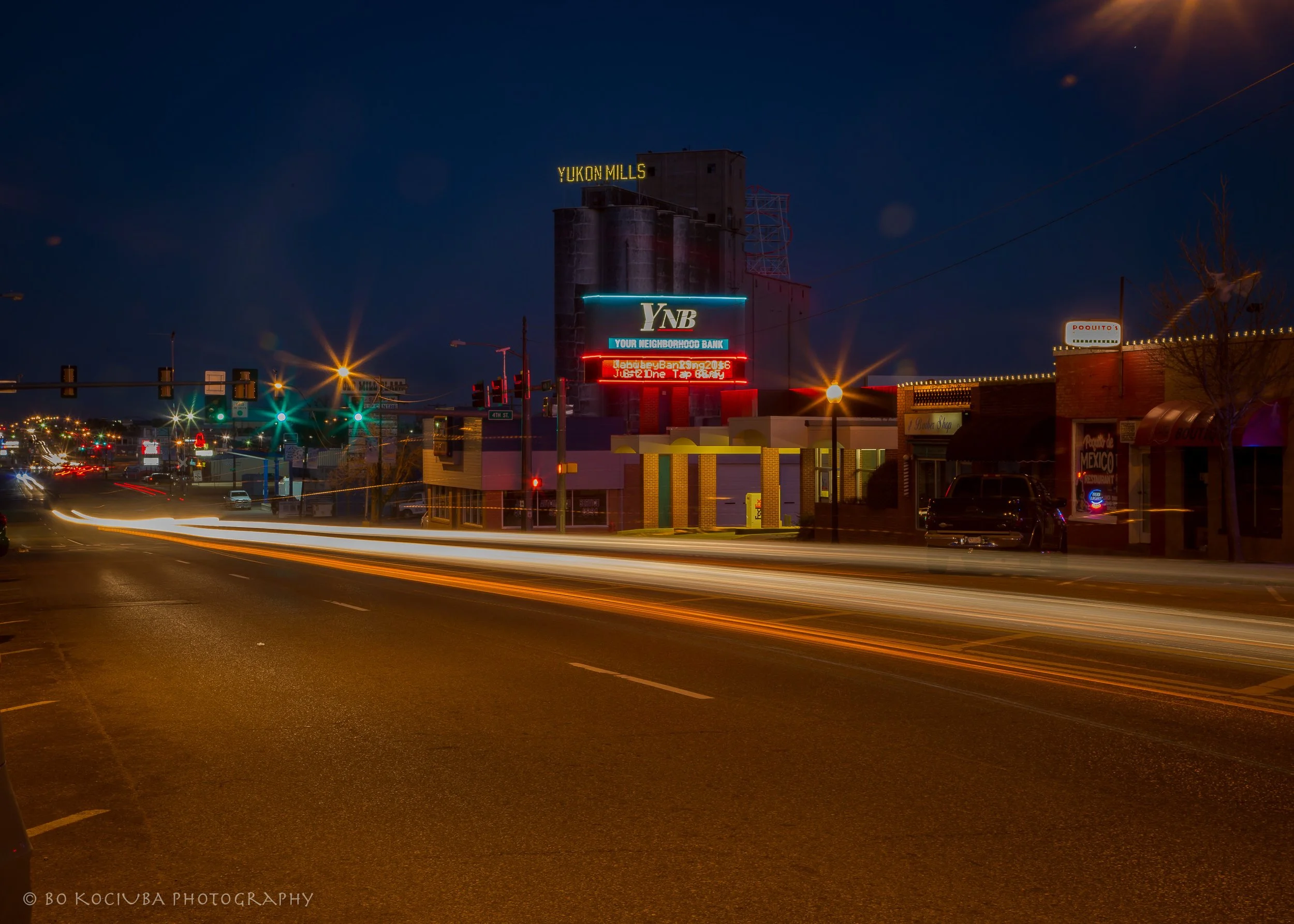 Historic Route 66 along Main Street in Yukon, Oklahoma, with illuminated business signs, storefronts, and light trails from passing vehicles at night.