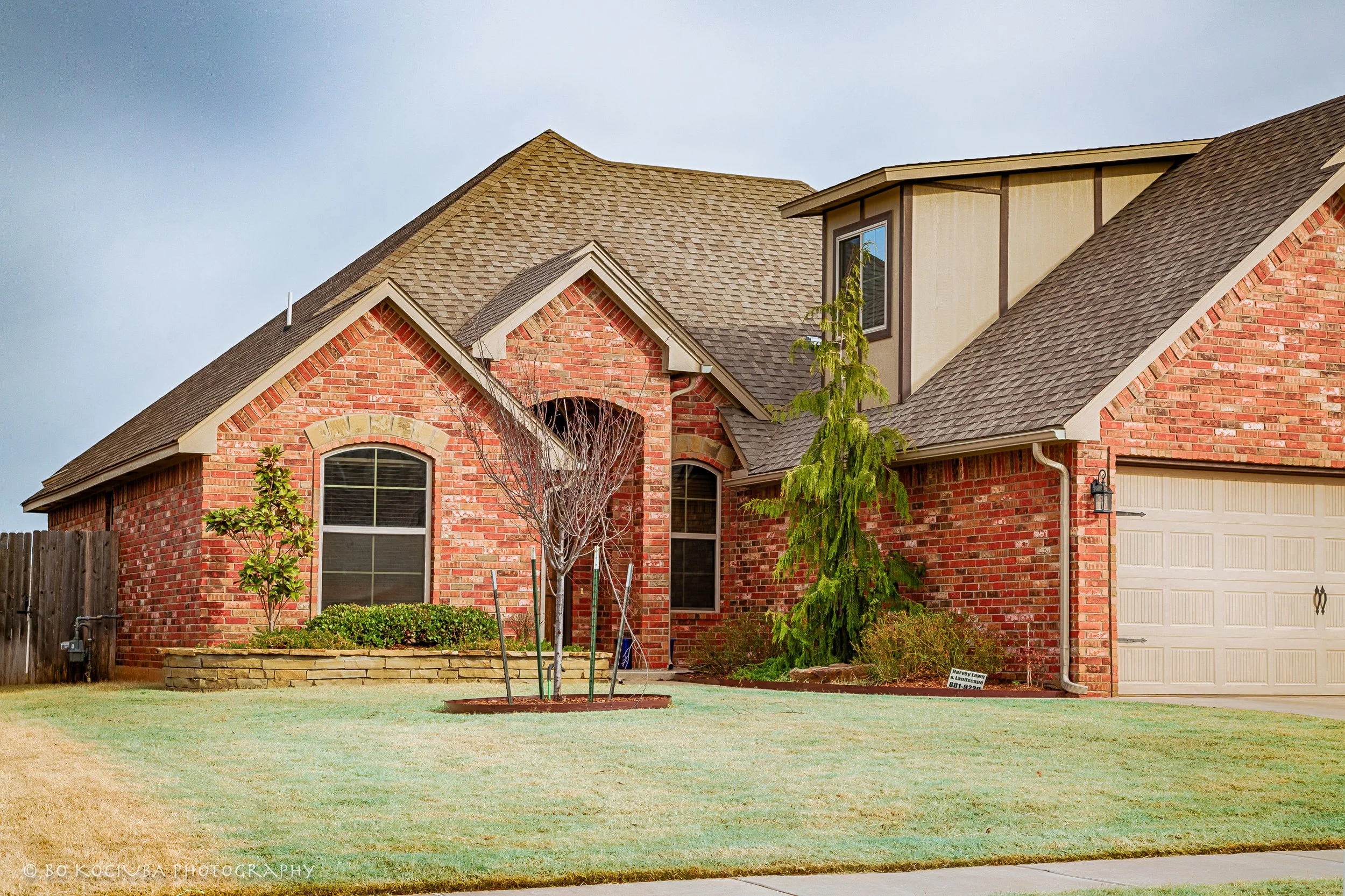 Red brick home in Chisholm Crossing, Yukon, Oklahoma, featuring multiple gabled rooflines, a covered front entry, attached garage, and landscaped shrubs.