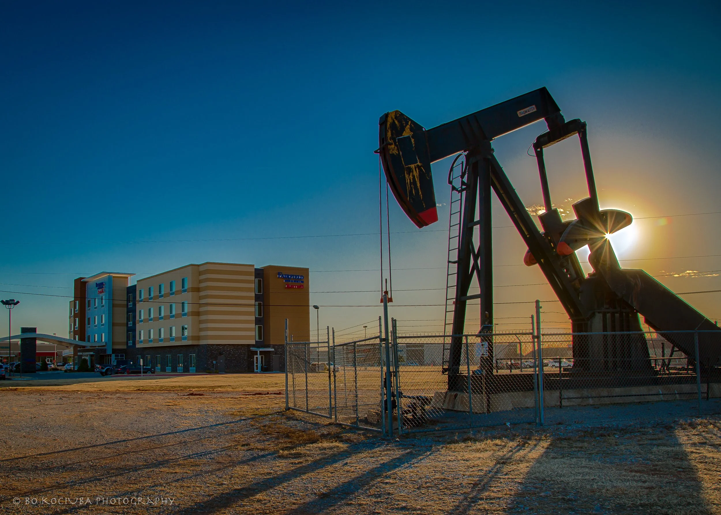 Oil pumpjack silhouetted against the setting sun in Yukon, Oklahoma, with a hotel and other commercial buildings in the background.