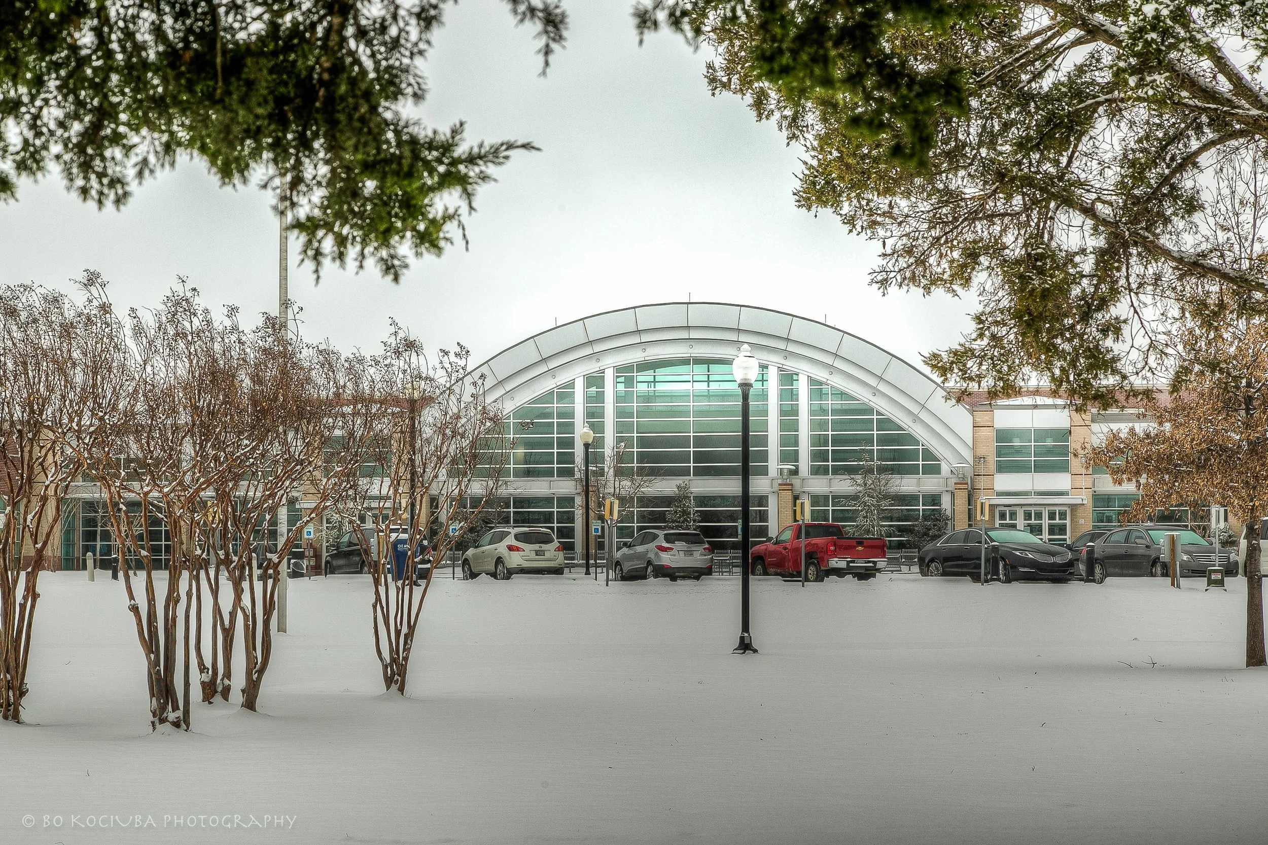 Community Center building with a large arched glass entrance in Mustang, Oklahoma, photographed after snowfall with trees, streetlights, and a parking area.
