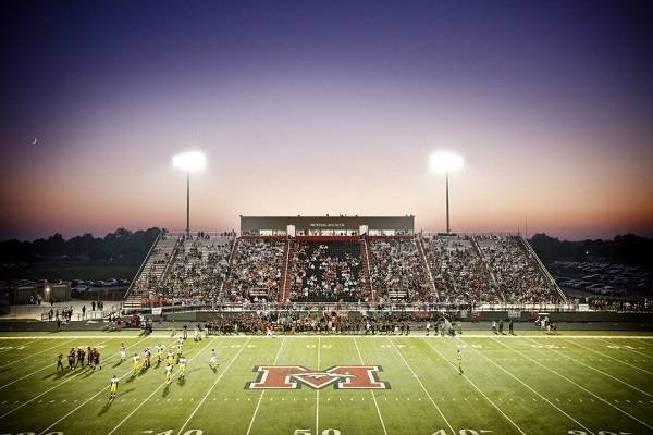 Mustang High School football stadium in Mustang, Oklahoma, during an evening game, with players on the field, packed stands, and stadium lights glowing at sunset.