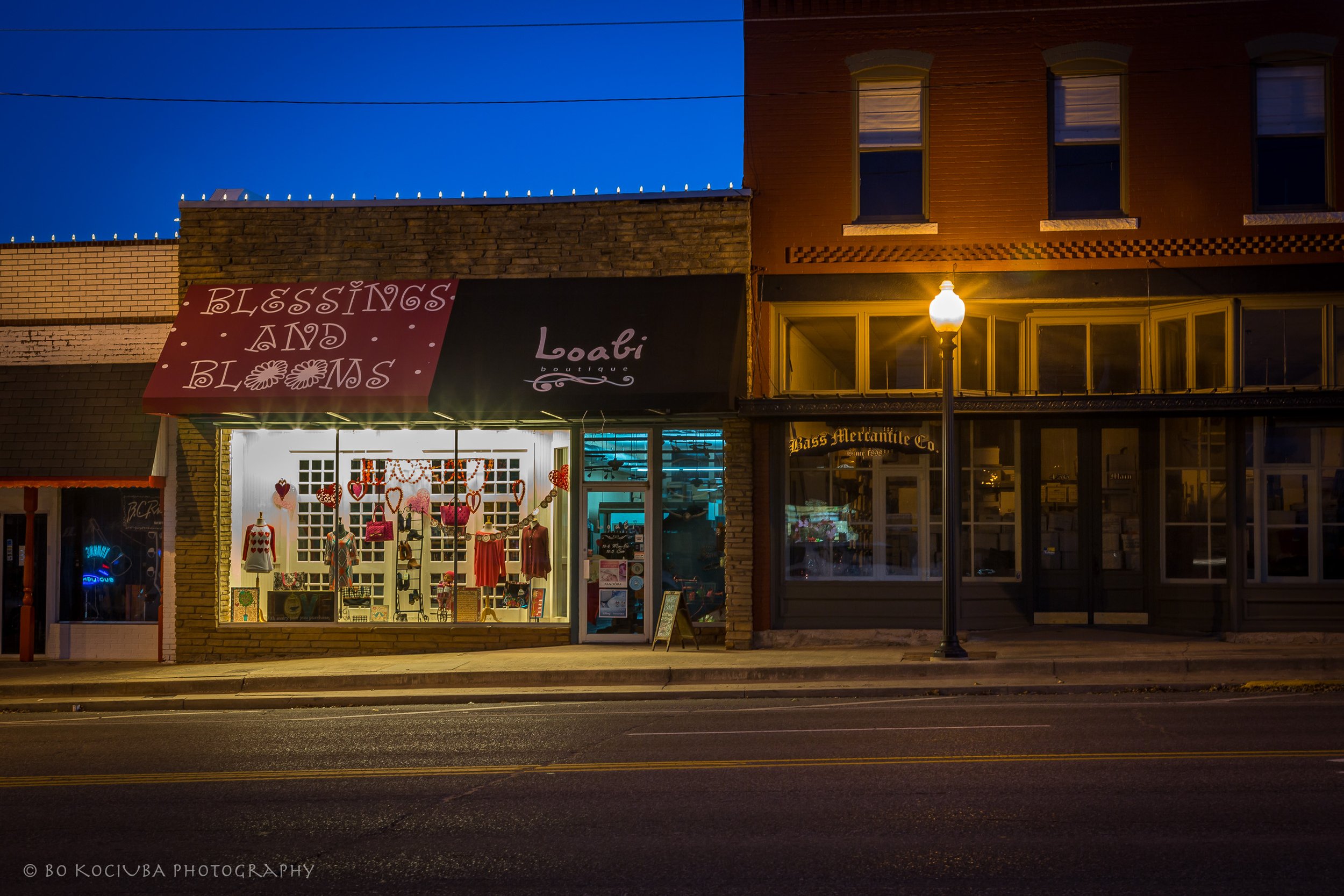 Historic Main Street in Yukon, Oklahoma, along Route 66 at night, featuring the illuminated storefronts of Blessings and Blooms and Loabi boutique.