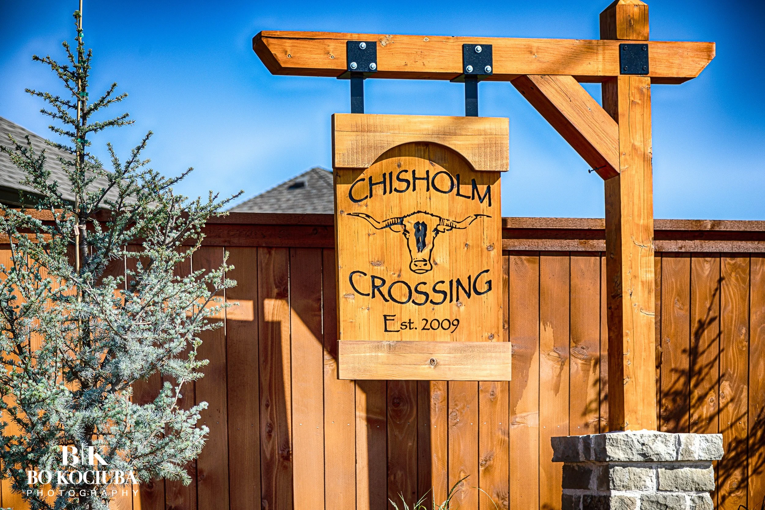 Wooden sign reading 'CHISHOLM CROSSING Est. 2009' attached to a wooden post, with a pine tree and a wooden fence in the background, under a clear blue sky.