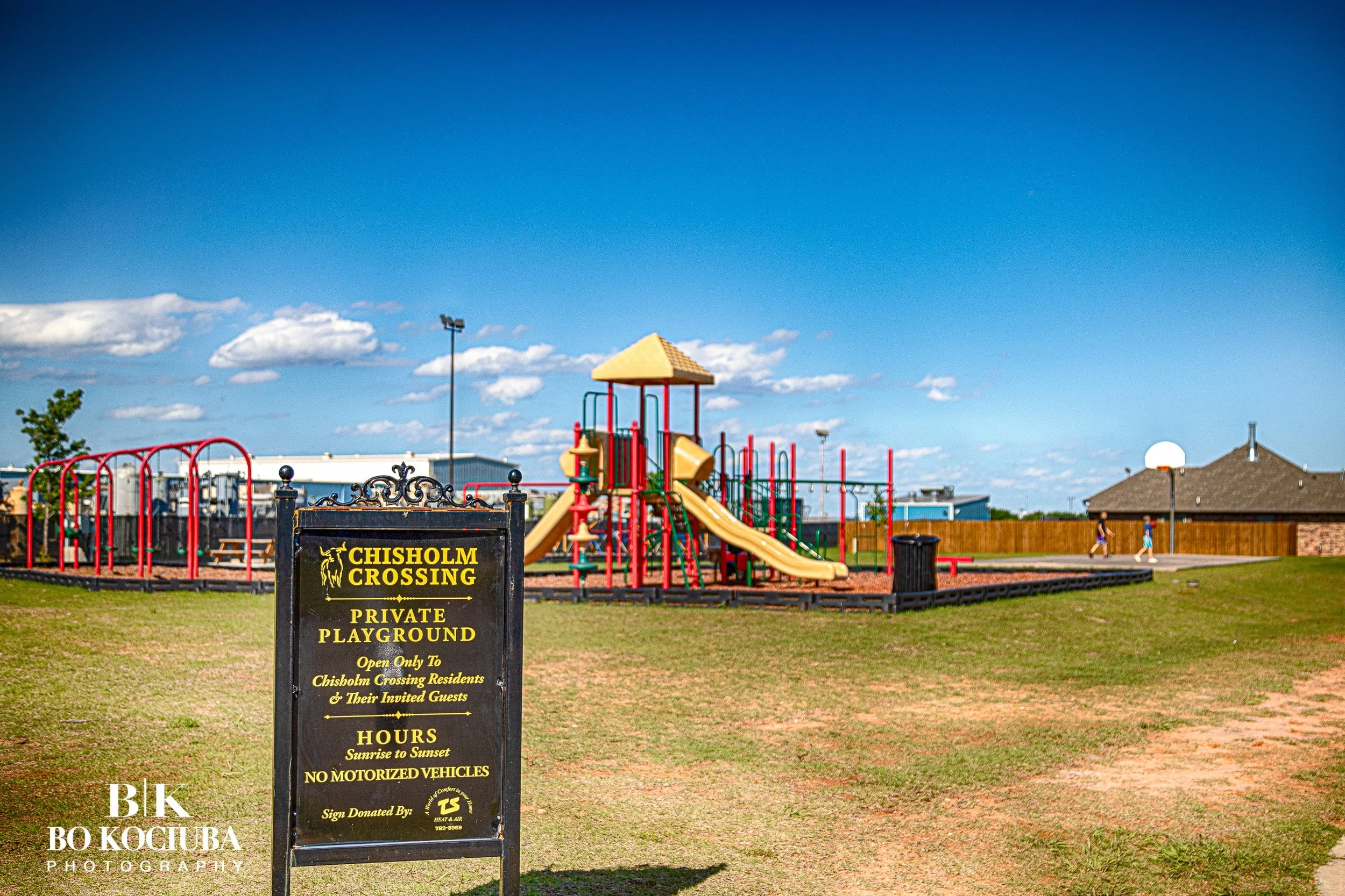 A playground with a slide, climbing structure, and jungle gym behind a black fence. A sign reads, 'Chisholm Crossing Private Playground.' The sky is blue with some clouds, and there are two children playing in the background.