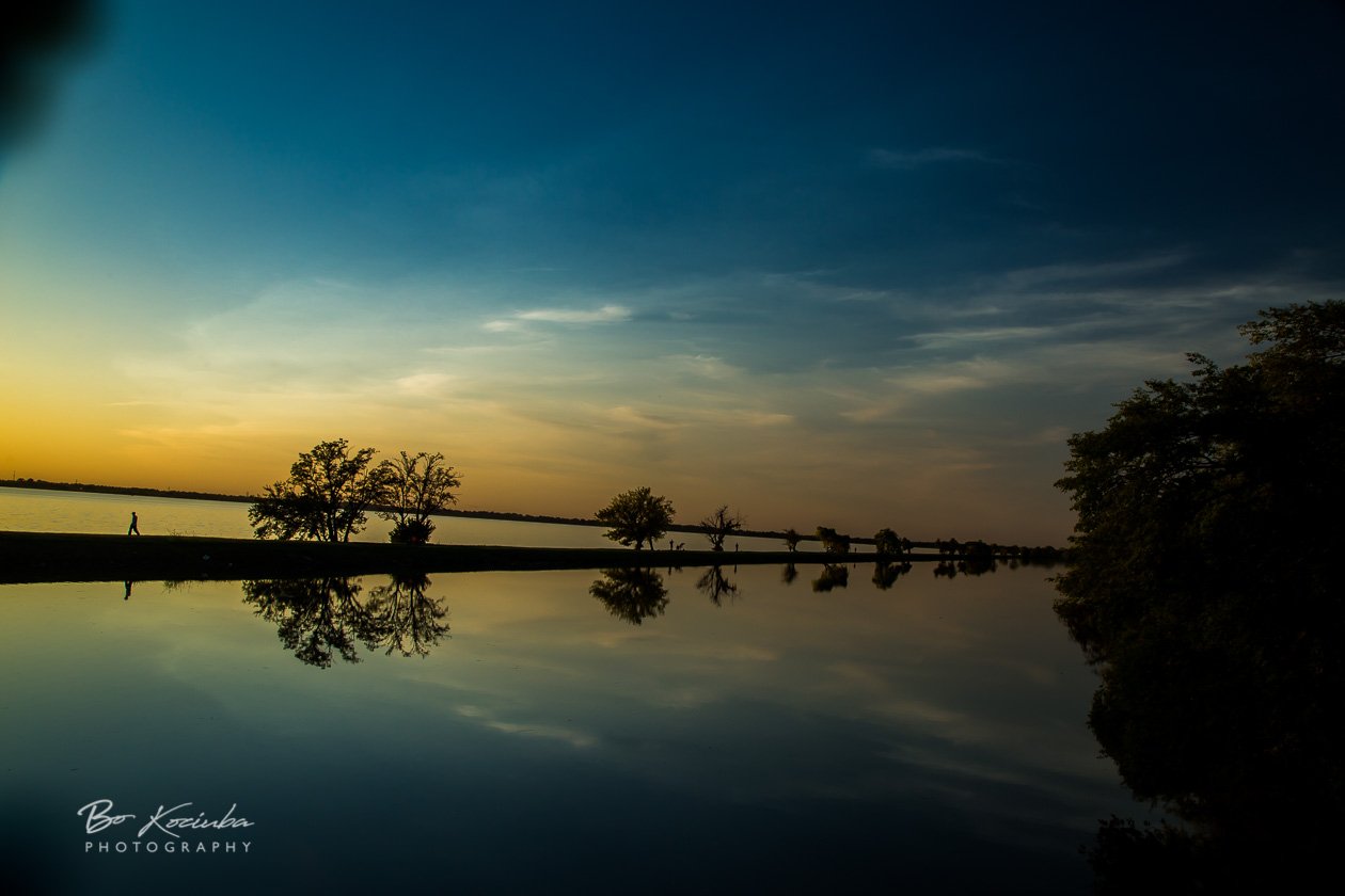 Sunset over Lake Overholser near Yukon, Oklahoma, with silhouetted trees along the shoreline and colorful evening light reflected across the calm water.