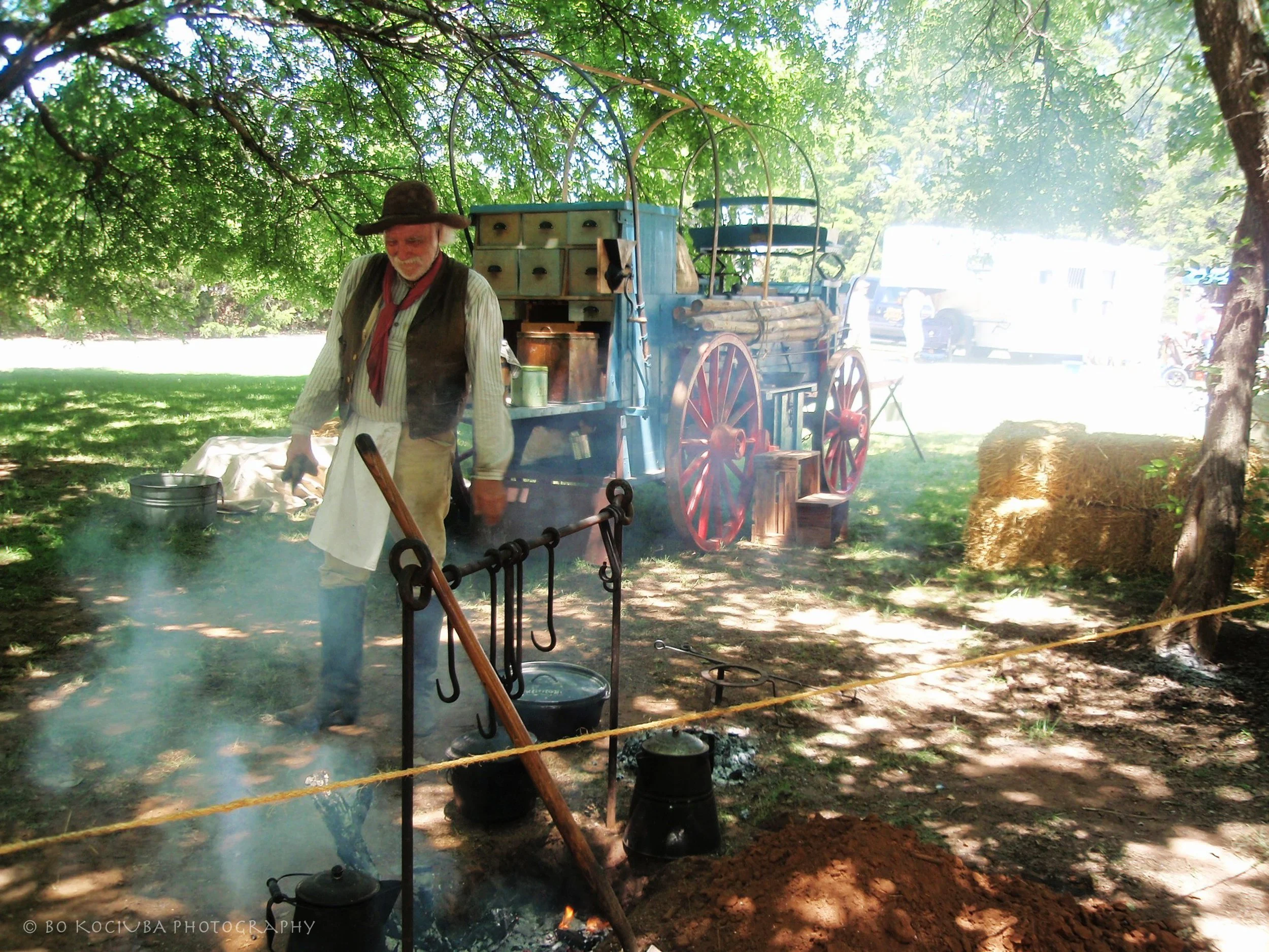 Historic chuckwagon cooking reenactment at the Chisholm Trail Festival in Yukon, Oklahoma, featuring a costumed participant preparing food beside a horse-drawn wagon and outdoor cooking equipment.