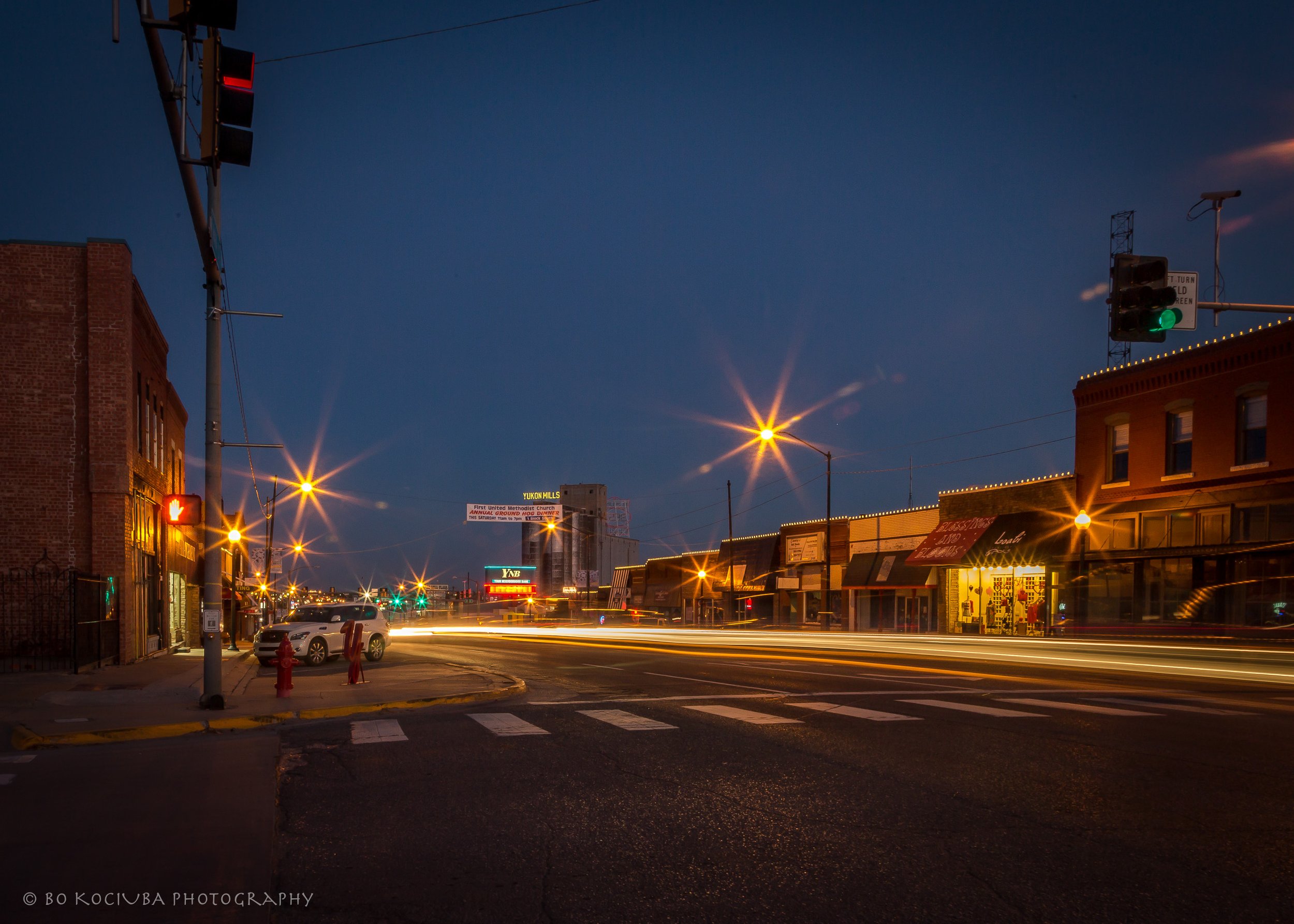 Historic Main Street along Route 66 in downtown Yukon, Oklahoma, illuminated by streetlights and storefronts, with vehicle light trails at dusk.