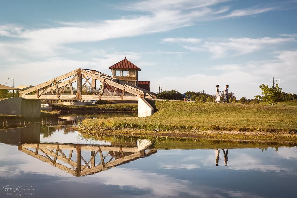 Lake Overholser Park near Yukon, Oklahoma, featuring a wooden pedestrian bridge over the water, a red-roofed pavilion, and visitors walking along the grassy shoreline.