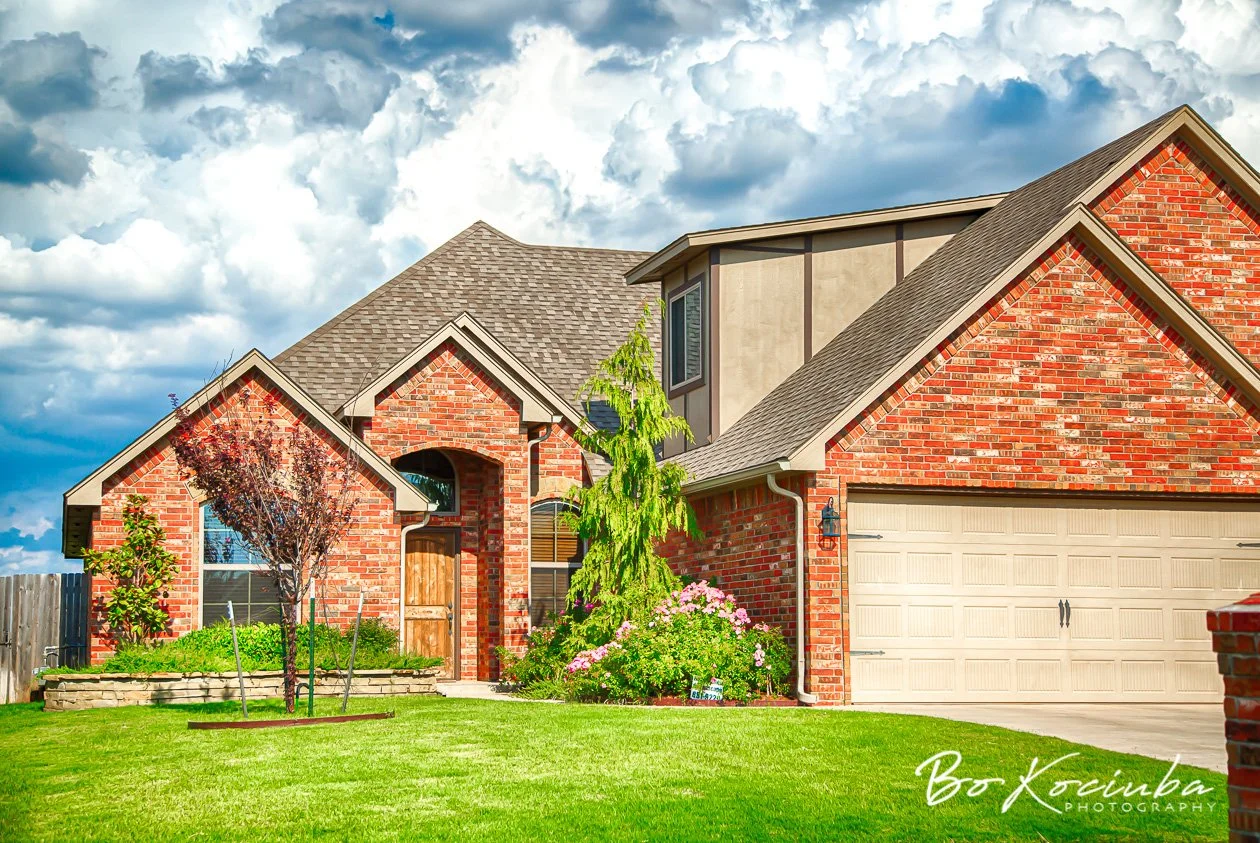 Brick homes in Chisholm Crossing, Yukon, Oklahoma, featuring gabled rooflines, green front lawns, flowering shrubs, and trees along the residential street.