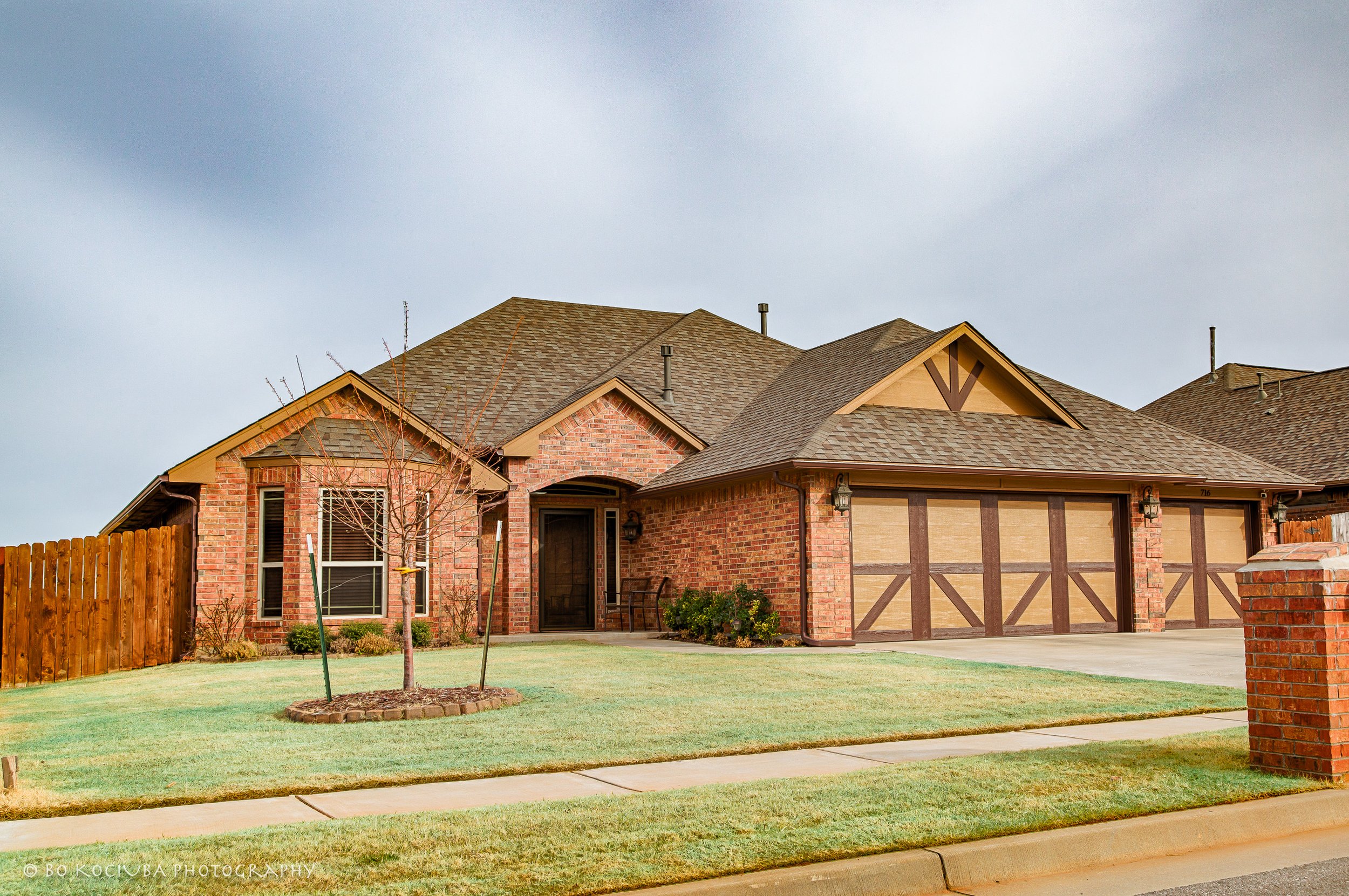 Brick home in Chisholm Crossing, Yukon, Oklahoma, featuring multiple gabled rooflines, decorative garage doors, a front lawn, and young trees.