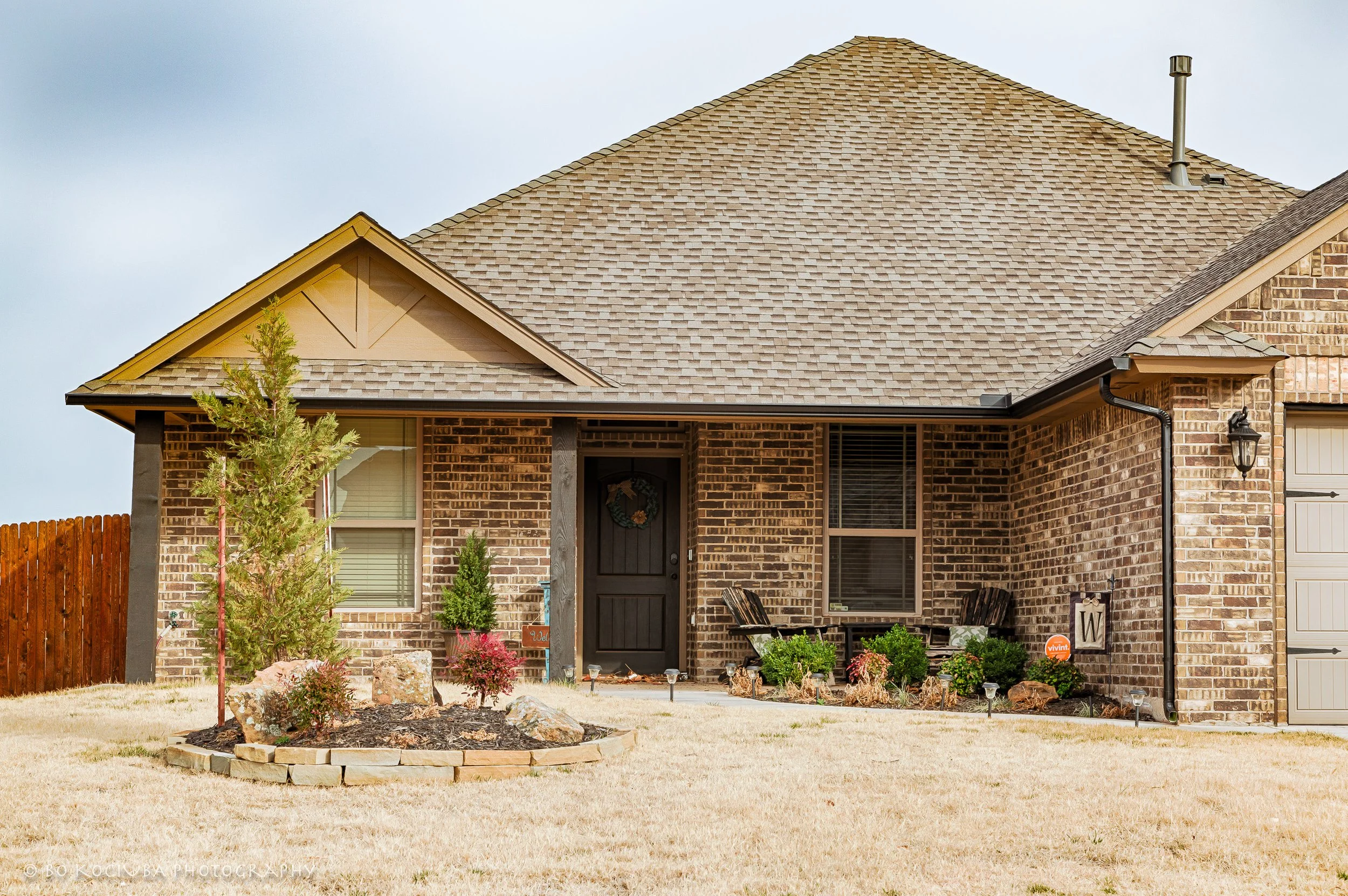 Brick home in Chisholm Crossing, Yukon, Oklahoma, featuring a covered front porch, gabled roof, young tree, and landscaped front garden.