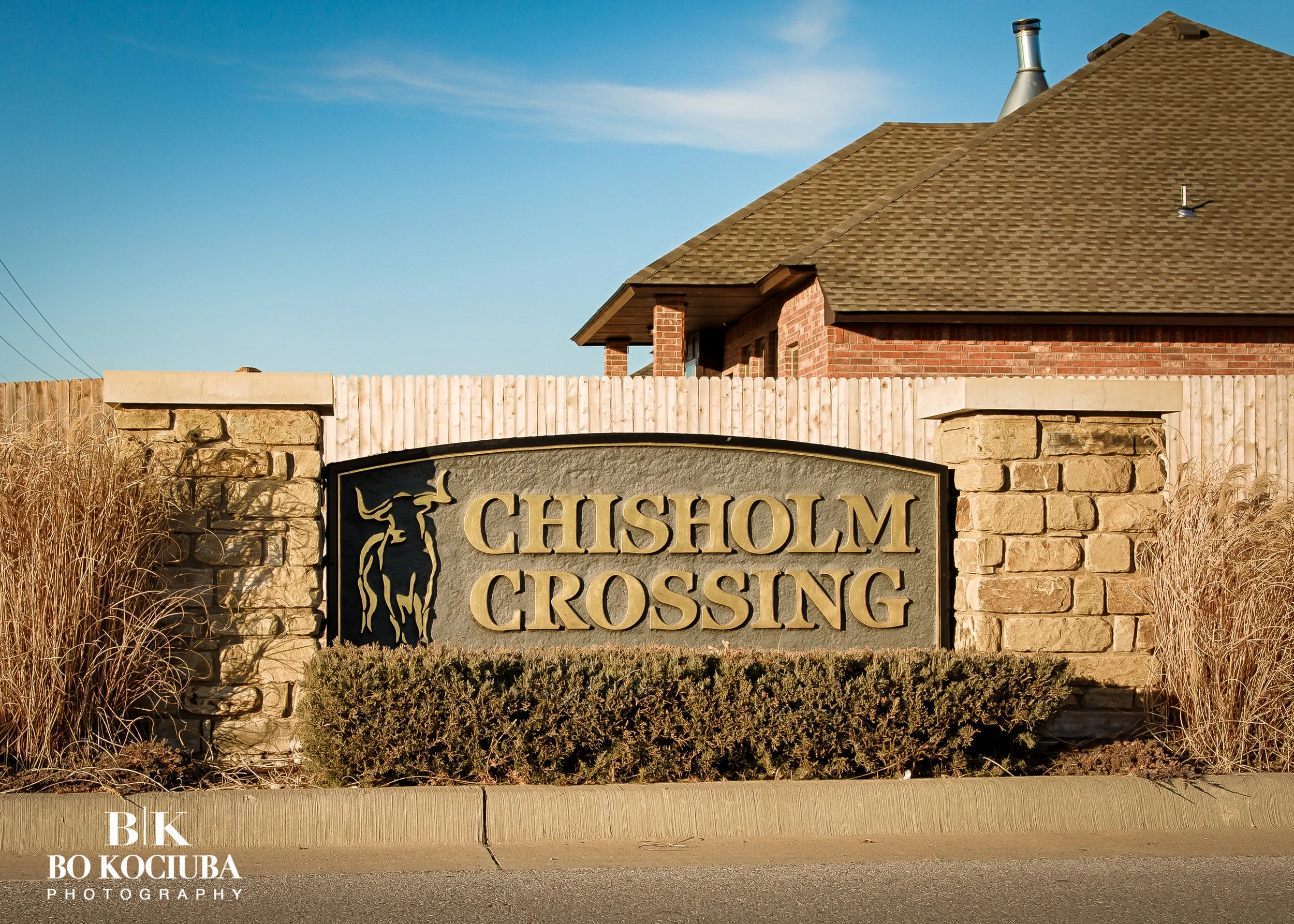 Stone and wood entrance monument for Chisholm Crossing in Yukon, Oklahoma, with a brick home visible behind the neighborhood sign.