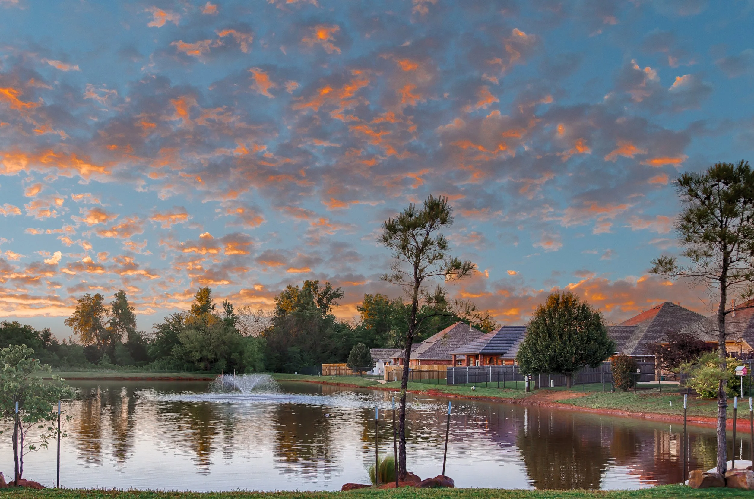 Neighborhood pond and fountain in Hunters Hill, Mustang, Oklahoma, surrounded by trees and residential homes beneath a colorful evening sky.
