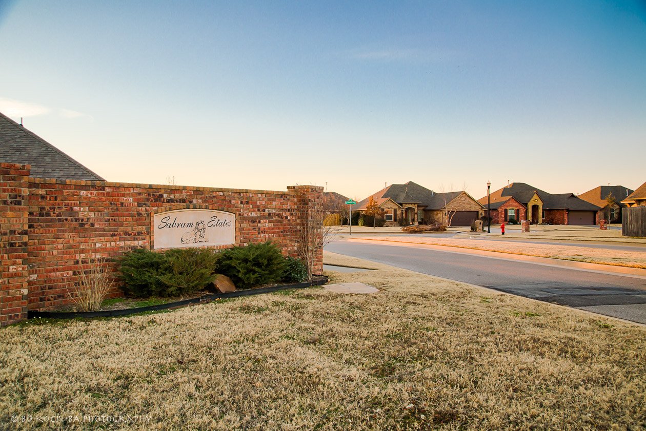 Brick entrance monument for Sabram Estates in Yukon, Oklahoma, with landscaped grounds, a residential street, and neighborhood homes in the background.